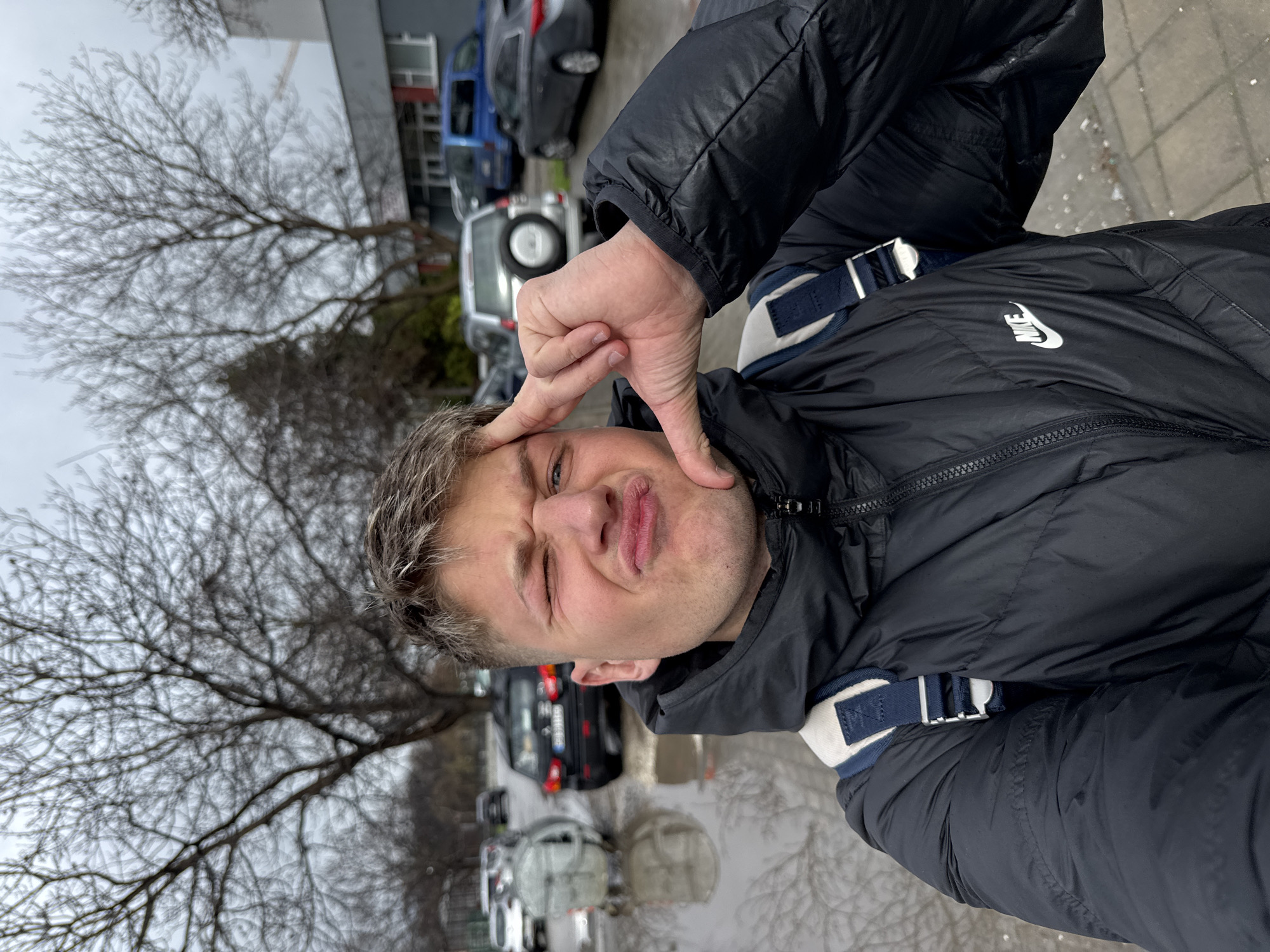 A man is standing outside in what appears to be a parking lot. He is wearing a black jacket with a white Nike logo on the chest and a strap across his shoulder. He is making a silly face and holding his hand up to his forehead. In the background, there are several cars and trees without leaves. The ground appears to be wet. It is not possible to determine the location in the city of Plovdiv, Bulgaria from the given information.