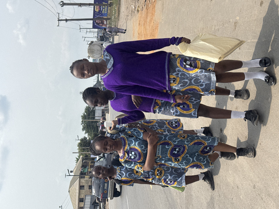Ogbaku, Nigeria. Four young females are depicted walking along the shoulder of a paved road under a bright sky with scattered clouds. The females are wearing school uniforms; the two leftmost individuals wear patterned short-sleeved dresses, while the two rightmost individuals wear purple long-sleeved sweaters over patterned skirts. The patterned fabric includes a logo with a book and laurel wreath, along with text partially visible as "SECOND POWERFUL" and "NWANA". The second female from the left has her arms crossed. The third female from the left holds a white cylindrical object in her right hand. The fourth female, positioned in the foreground to the right, carries a light-colored plastic bag in her right hand and is looking directly towards the viewer. Footwear visible includes dark athletic shoes, black flats, and white socks. In the background, a dark SUV is parked on the road, and various buildings, utility poles with overhead lines, and commercial signage are visible. Another individual walks in the distance on the right side of the road.