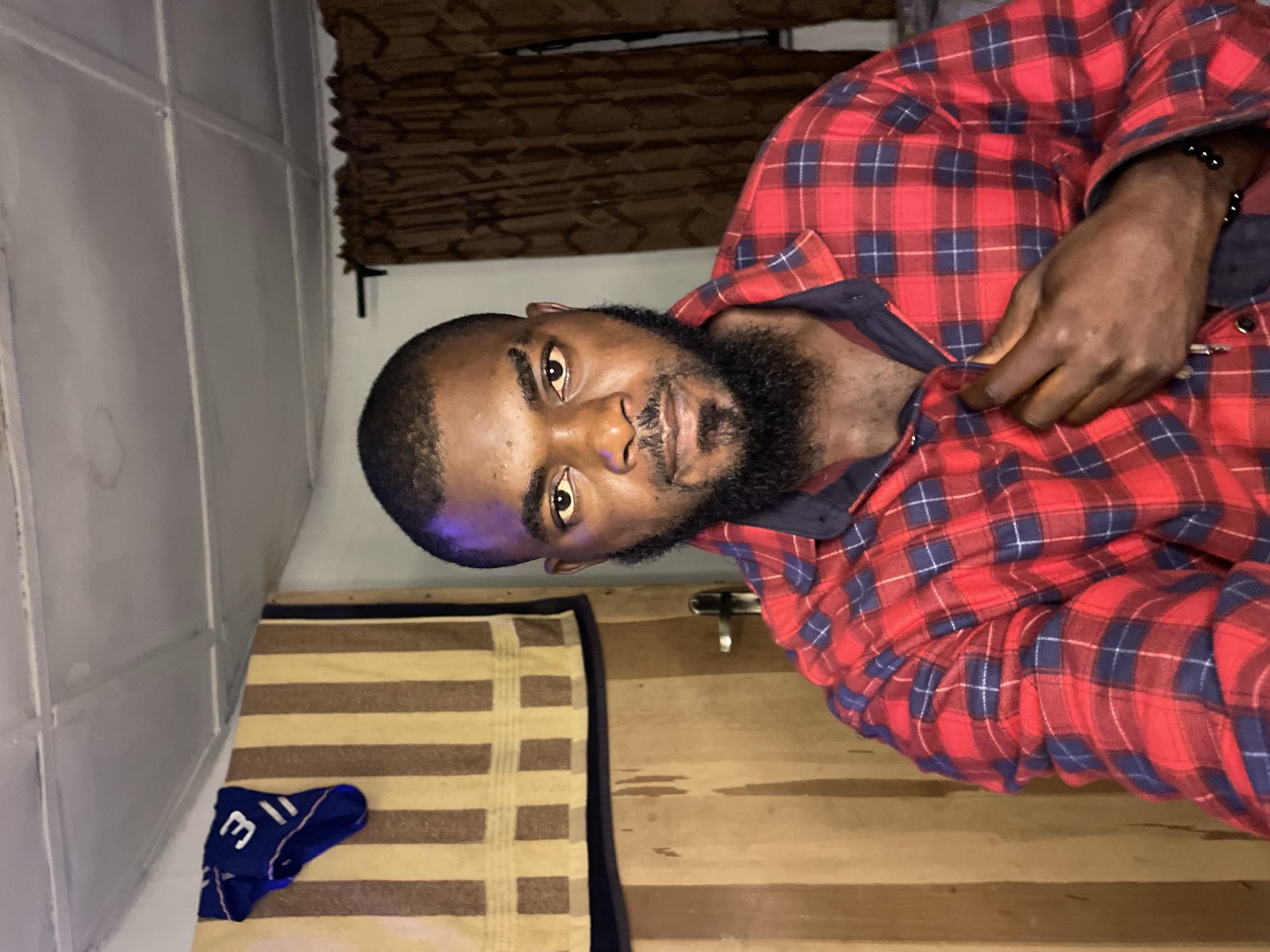 A waist-up, medium shot shows a man with dark skin, a beard, and short hair, looking directly at the camera. He is wearing a red and navy blue plaid shirt, partially buttoned. His right hand is on the shirt, near the buttons. The man is positioned in the center of the frame, with a wooden door and a striped curtain to his left, and a brown curtain behind him to the right. A blue garment with the number "31" is visible hanging on the wall in the upper left corner. The ceiling appears to be made of suspended tiles, with some visible staining. The lighting suggests an indoor environment, possibly at night. The scene is not specifically identifiable as being within the city of Jos, Nigeria, based solely on the visual information provided.