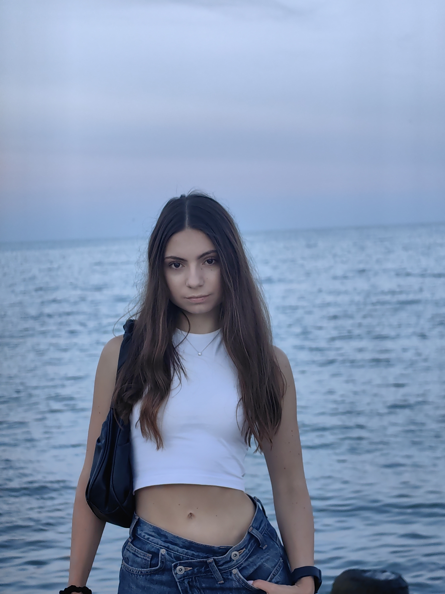 A young woman with long, dark brown hair stands facing the camera with a neutral expression. She is wearing a white crop top and denim jeans. A black shoulder bag is slung across her left side. She is standing on what appears to be a pier or rocky shoreline, with the ocean and a pale blue sky in the background. The lighting suggests it is dusk or early morning.