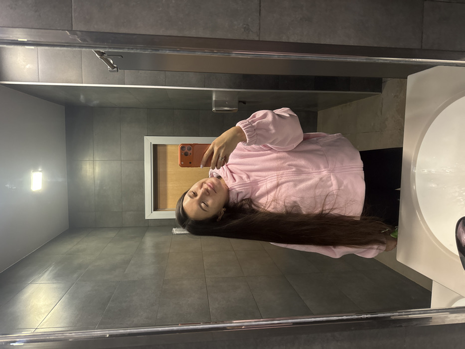 A female individual is captured in a mirror selfie within a public restroom in Obninsk, Russia. The subject has long, dark brown hair falling over her left shoulder and chest, and wears a light pink, full-zip, possibly fleece jacket over black pants. Her head is tilted slightly to her left, with eyes partially closed and a subtle smile. She holds an orange-cased smartphone in her right hand, positioned to photograph her reflection. A ring is visible on her right ring finger. The restroom features dark gray rectangular wall tiles, a white rectangular sink with a chrome faucet in the foreground, and a light-colored wooden door with a silver-toned frame visible in the background. A metallic fixture, potentially a hand dryer or paper towel dispenser, is mounted on the wall to the right of the reflection. A rectangular overhead light illuminates the space. The floor is tiled with lighter-colored tiles.