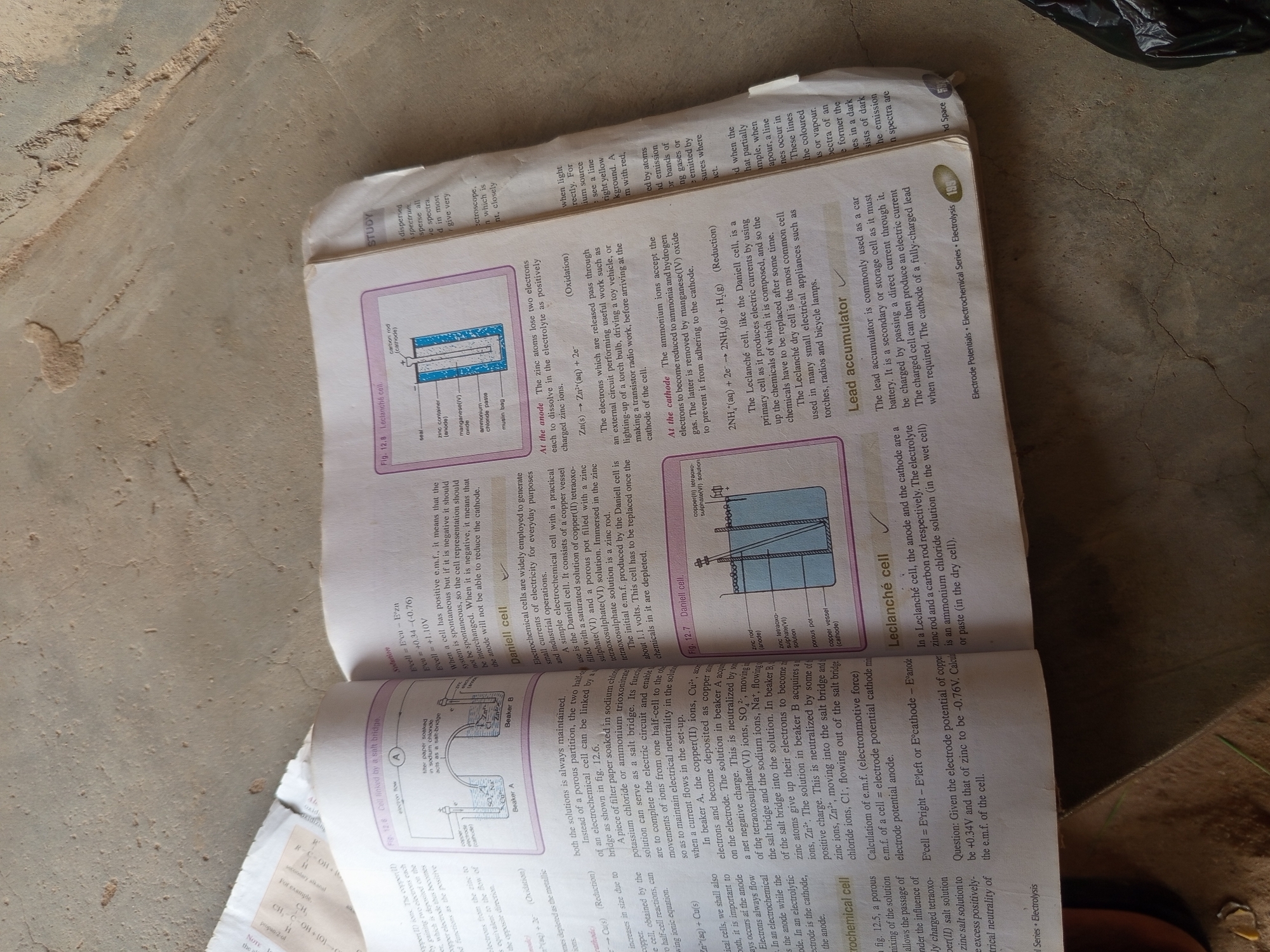 The image is of an open textbook, likely a science or chemistry textbook, displaying diagrams and text related to electrochemical cells, specifically the Daniell cell and Leclanché cell.
The textbook is lying flat on a textured, possibly concrete or stone surface, with a light-colored, rough, and uneven texture. There are also shadows present on the surface, suggesting overhead lighting. The overall environment appears to be indoors, possibly a study area or a classroom. The presence of a dark, crumpled object in the lower right corner suggests it might be a bag or some other personal item.
There are no people or identifiable landmarks in the image, and therefore no information can be provided regarding the city of Jalingo, Nigeria, or any specific location within it. The focus is entirely on the educational content within the textbook.