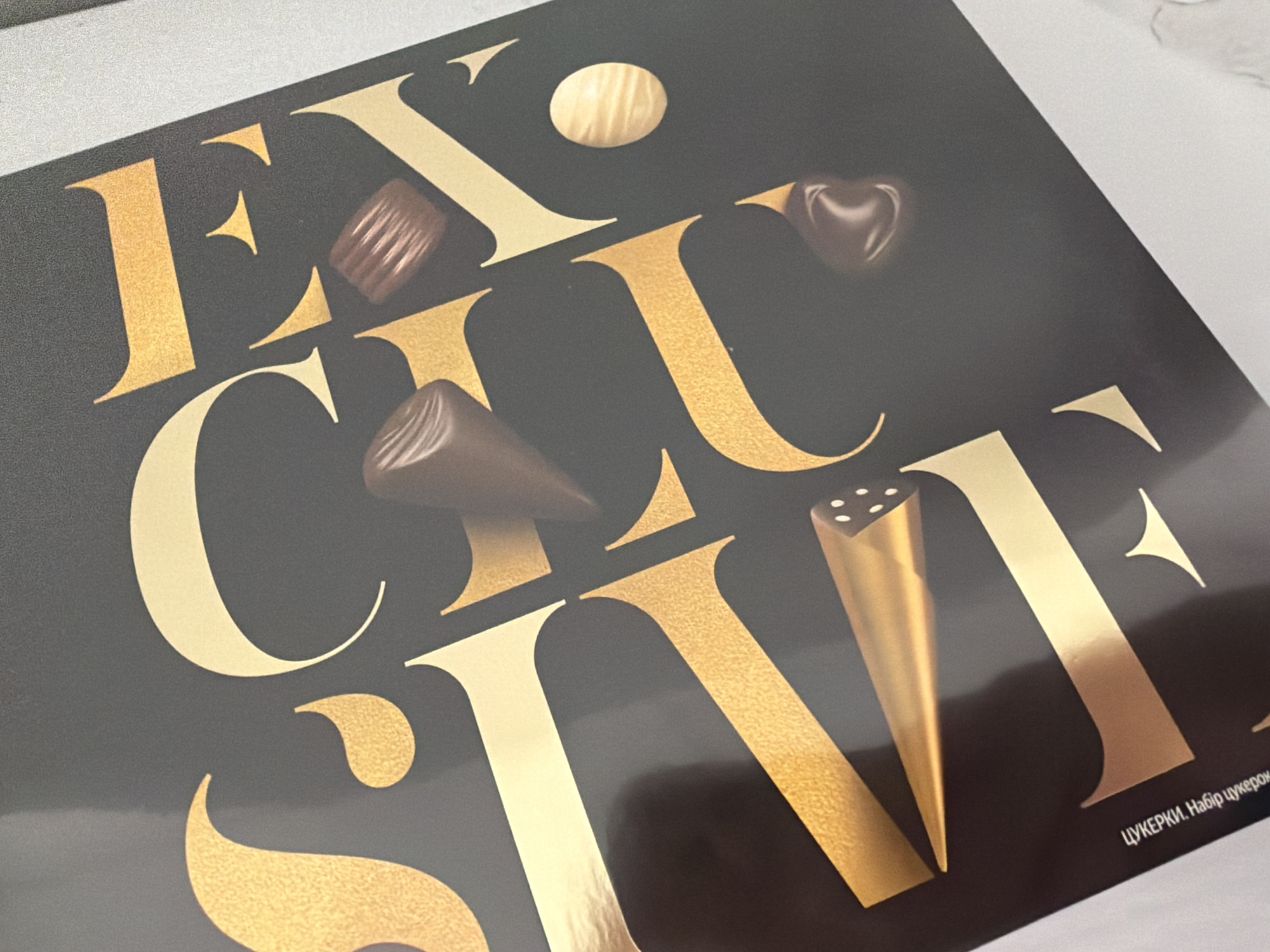 The image shows a close-up of a dark background with large, stylized gold and cream-colored letters that appear to spell out a word. There are several chocolates scattered on the surface, with some partially obscuring the letters. In the bottom left corner, there is text in a smaller font that reads "ЦУКЕРКИ. Набір цукерок", which translates to "CANDIES. Set of candies" in Ukrainian. The overall aesthetic suggests a product packaging or advertisement. There is no indication of the city of Kyiv or Ukraine in the image itself.