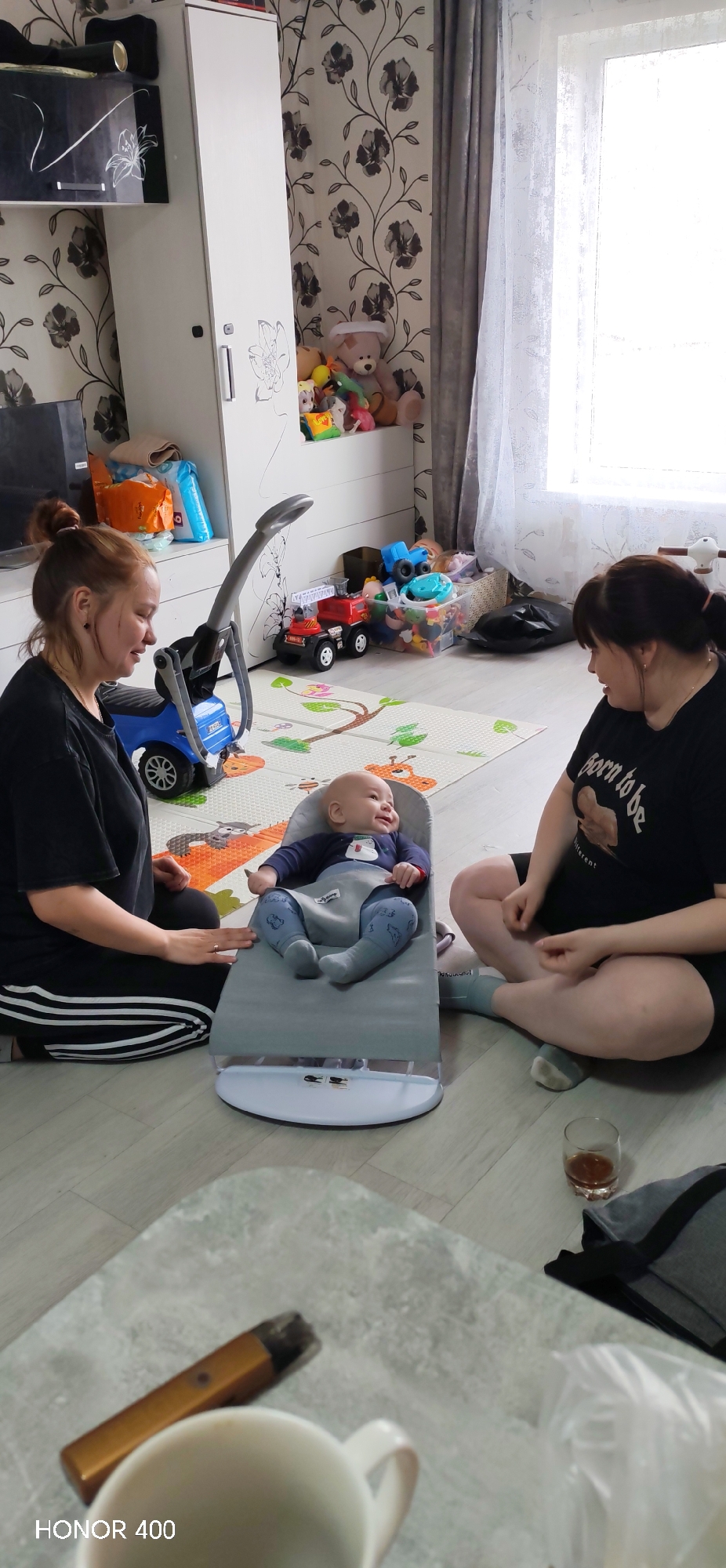 An indoor scene captured in Пушкари, Russia, shows two women and an infant interacting on a light-colored wooden floor. The room features patterned wallpaper with dark floral motifs and a large window with sheer white curtains, through which bright daylight streams.
In the center, an infant in a blue outfit lies smiling in a gray baby bouncer. To the left, a woman with reddish-brown hair tied back sits cross-legged, wearing a black top and black track pants with white stripes, looking down at the baby with a gentle expression. To the right, another woman with dark hair tied back also sits cross-legged, wearing a black t-shirt with the visible text "born to be" followed by an obscured word, looking towards the baby. Between the women and beneath the bouncer is a colorful playmat with animal designs.
Various children's toys, including a blue ride-on vehicle, red fire truck, and plastic bins filled with other toys and plush animals, are scattered on the floor and stored on shelves of a white cabinet. The cabinet itself has decorative floral patterns. In the foreground, a white ceramic cup and a gold-colored vape pen rest on a textured, light-colored surface, likely a table. A glass containing an amber liquid is visible on the floor near the right woman. The bottom left corner of the image bears a "HONOR 400" watermark. The overall atmosphere appears relaxed and domestic.