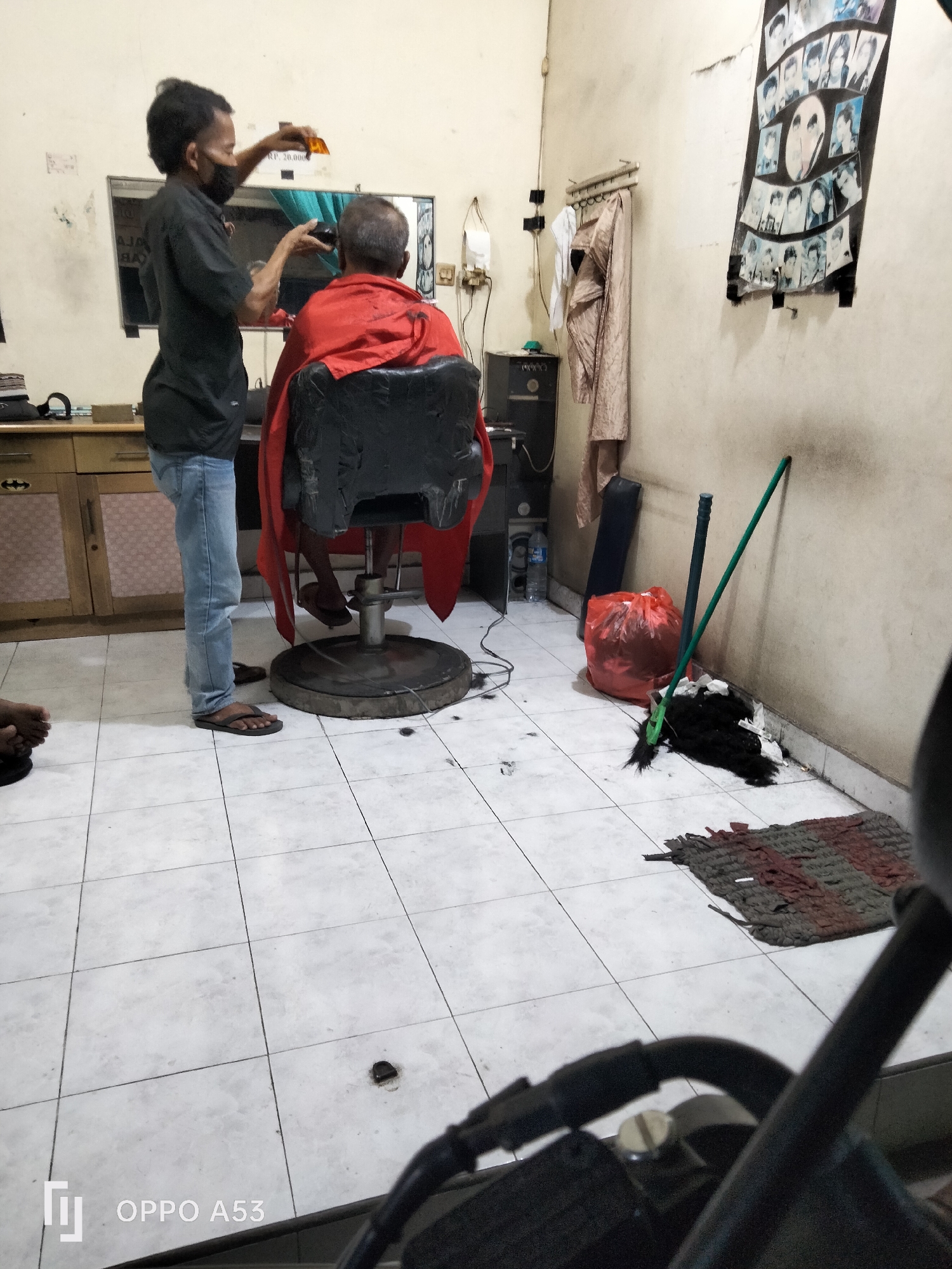 The image depicts the interior of a barbershop, likely located in Denpasar, Indonesia. A barber, wearing a black shirt and blue jeans, is in the process of cutting the hair of a client. The client is seated in a black barber chair, draped in a red cape. The barber is holding a pair of scissors and a comb. Behind the client, a mirror reflects the scene. To the left of the barber, a wooden cabinet with glass doors is visible. On the wall to the right, a collage of photographs is displayed. In the foreground, a red trash bag and cleaning supplies, including a broom and mop, are present on the tiled floor. The overall environment appears to be a modest, functional barbershop.