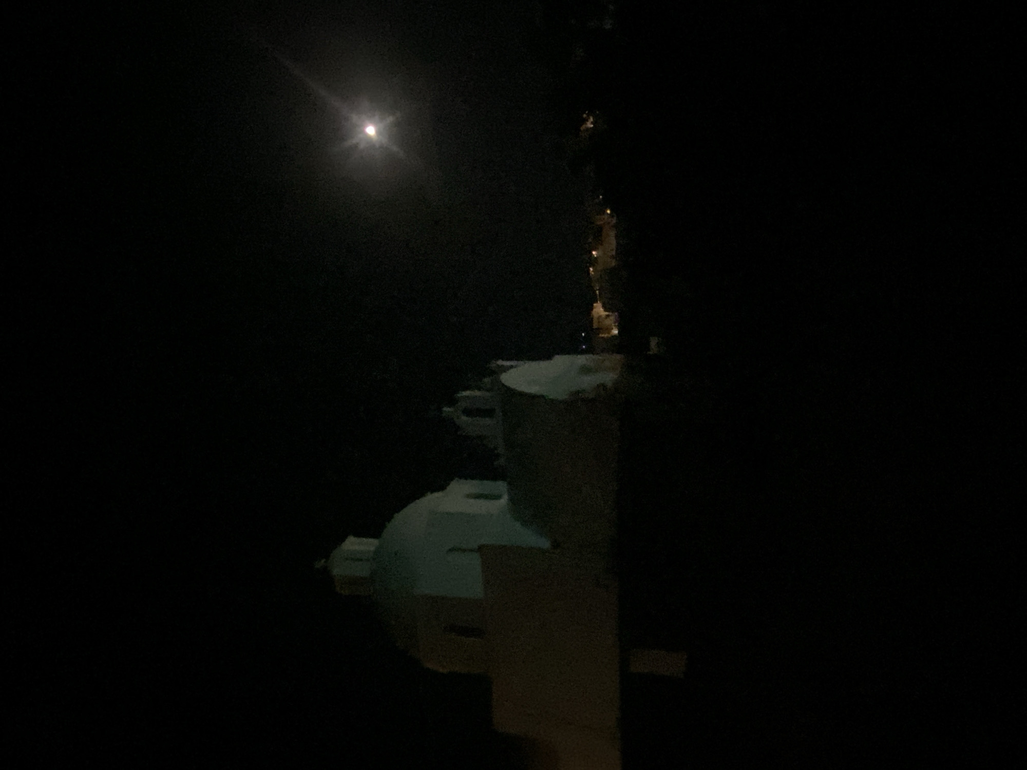This is a night-time photograph of a white building with a dome, likely a church, in the city of Thira Municipal Unit, Greece. The building is illuminated and stands out against the dark sky. In the upper right portion of the image, the moon is visible, casting a bright glow and a lens flare. The background shows a faint outline of other buildings and lights, suggesting a populated area. The overall scene is dark and atmospheric, with the moon and the illuminated building as the primary focal points.
