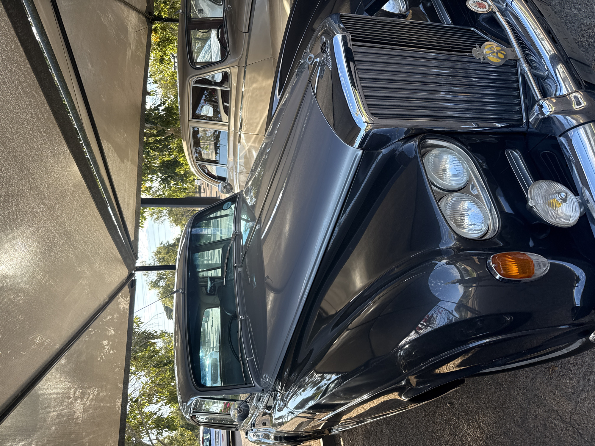 An eye-level, medium shot shows two vintage cars parked under a shelter in Stellenbosch Nu, South Africa. The car in the foreground is a dark, possibly black, vintage vehicle. The hood is highly polished, reflecting light and the surrounding environment. A chrome grill with vertical slats is visible at the front of the car. Two large, circular headlights are present, along with a smaller side light. An orange turn signal is also visible.
The second car, parked behind the first, appears to be a lighter color, possibly beige or cream. Only part of this car is visible.
The cars are parked under a shaded structure, possibly a car port. Support beams are visible. Trees and vegetation are visible in the background. The pavement can be seen beneath the vehicles.
