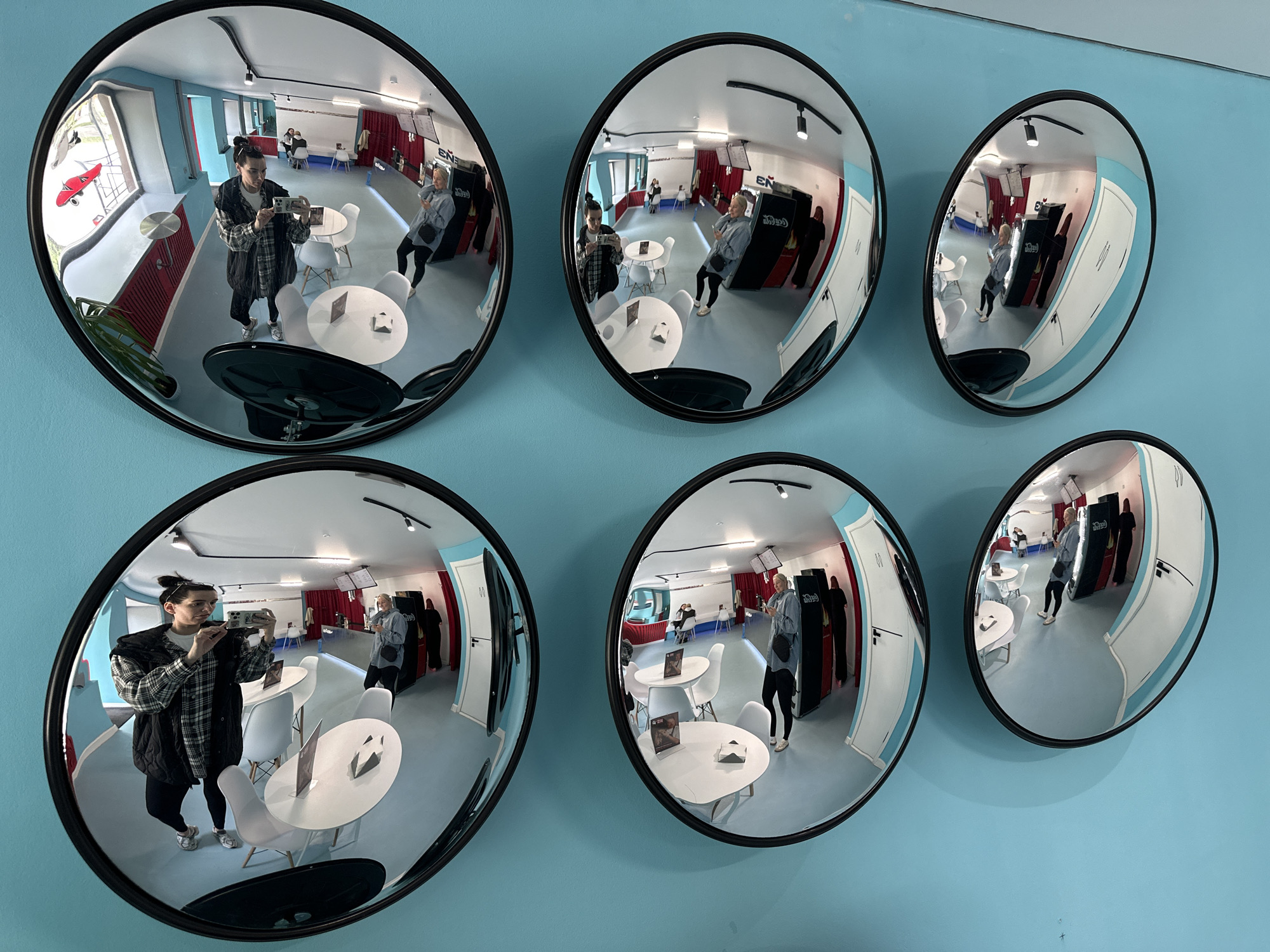 The image depicts a row of convex mirrors mounted on a bright blue wall, likely in a public space within the city of Chaikovsky, Russia. The mirrors reflect the interior of what appears to be a cafe or lounge area.
In the foreground, a person wearing a plaid shirt and a dark vest is taking a selfie in the largest mirror on the left, holding a smartphone. They are wearing dark leggings and white sneakers. In this same mirror's reflection, another person in a light blue shirt and dark pants is visible standing near a counter.
The reflections in the mirrors show a bright, modern interior with light blue walls and light-colored flooring. Several small, round white tables with matching chairs are arranged throughout the space. In some reflections, other patrons can be seen sitting at tables or standing near a counter. Behind the counter, there are shelves with items and what look like vending machines or display cases. Red curtains are visible in the background of some reflections, adding a touch of color. The overall impression is a casual, contemporary establishment.