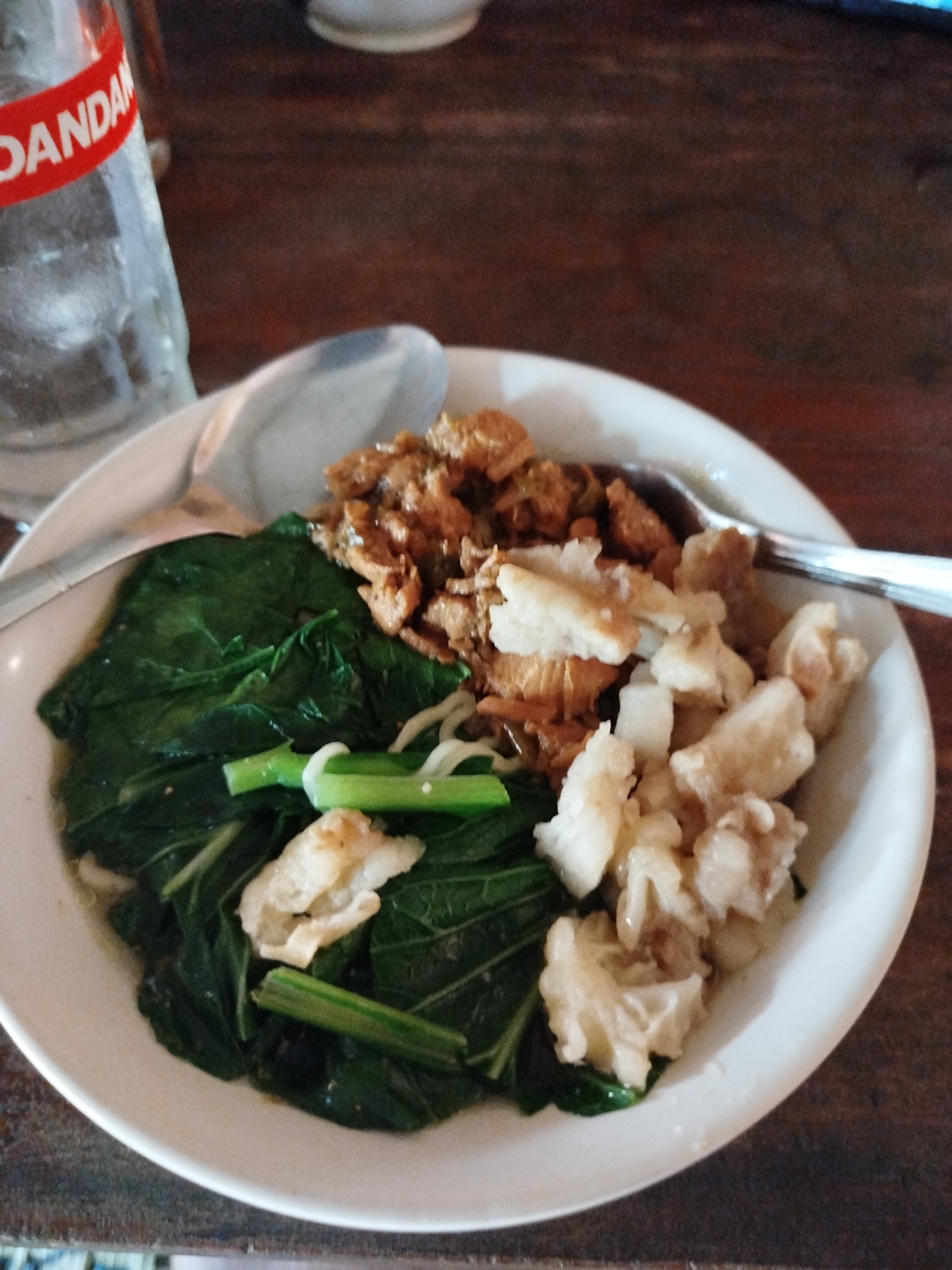 A bowl of food is presented on a wooden table, with a partially visible glass bottle of Dandanan drink to the left. The bowl contains noodles topped with shredded chicken or meat, a portion of dark green leafy vegetables, and several pieces of what appear to be fried chicken skin or some other crispy topping. A metal spoon rests in the bowl. The setting is likely a casual eatery in Pojok, Indonesia, given the brand of the beverage.