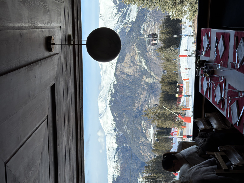 The image depicts an indoor scene looking out a large window onto a snowy alpine landscape in Courchevel, France.
**Interior:** In the foreground and left mid-ground, a dining area is visible. A long, rectangular wooden table is set with at least six place settings, each featuring a red placemat, a folded dark napkin, a knife, a fork, and a clear glass. Small bottles, likely condiments, and a dark decorative item are positioned in the table's center. Dark wooden chairs are partially visible. Two individuals are seated with their backs to the camera on the left; one wears glasses and has dark hair tied back, while the other is largely obscured, wearing a light-colored garment. The ceiling above consists of dark wooden panels, from which a spherical dark pendant lamp hangs in the upper right portion of the indoor frame.
**Exterior:** Beyond the window, a panoramic view of snow-covered mountains under a blue sky with scattered clouds dominates the background. Dark green evergreen trees sparsely cover the lower and middle mountain slopes. In the mid-ground, closer to the building, a groomed ski slope is active with several small figures of skiers and snowboarders. Red safety netting and white pole barriers delineate sections of the slope. A gondola lift system traverses the scene, with multiple dark-colored cabins visible, one of which prominently displays a white graphic on its side. The lighting suggests a bright, sunny day.