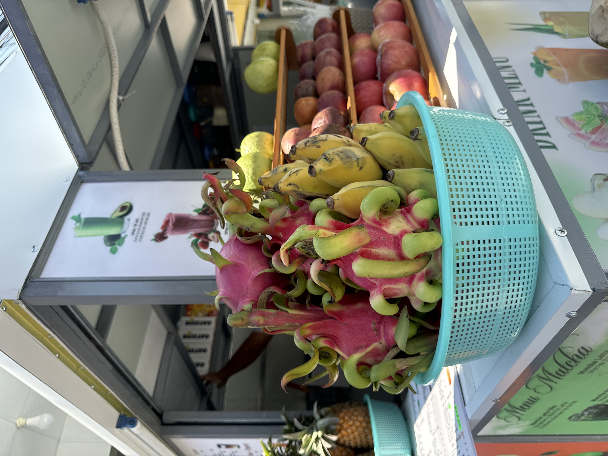 The image captures a fruit stand in Mũi Né, Vietnam. A blue plastic basket overflows with dragon fruit and bananas. Behind the basket are other fruits, including guavas, peaches, and apples, arranged on shelves. The stand is situated within a structure with metal framing. Printed signs displaying smoothie and drink menus are visible. A pineapple is present below the basket. An arm can be seen reaching into the stand.