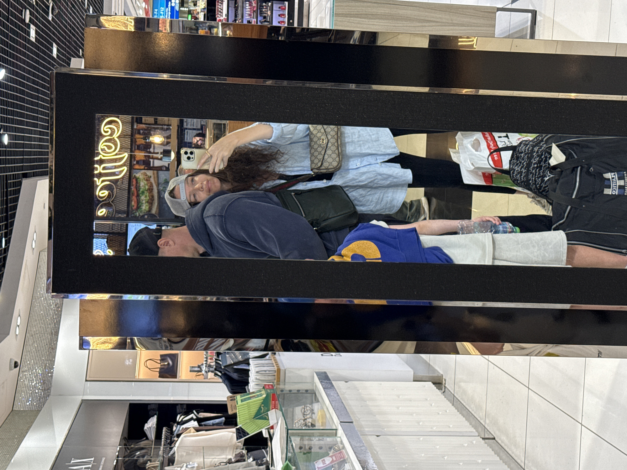 A woman, wearing a grey baseball cap and a light blue, unbuttoned shirt over a black top, takes a selfie in a mirror. She is holding a smartphone in her right hand and a beige Gucci crossbody bag with a gold logo pattern is slung across her body. To her left, a man wearing a dark blue hoodie and a black baseball cap is visible, his shoulder facing the camera. To the left of the man and woman, a child in a blue and yellow shirt is also partially visible.
The scene appears to be inside a retail establishment, possibly an airport duty-free shop given the overhead lighting and display shelves. In the background, illuminated signs for "coffee" are visible, along with displays of food items. To the far left of the image, racks of clothing and accessories are displayed in a shop with an "AX" logo. Various shopping bags and luggage are placed on the floor in the foreground. The overall environment suggests a commercial setting. The image is taken from a low angle, with the mirror creating a reflective surface that captures the subjects.