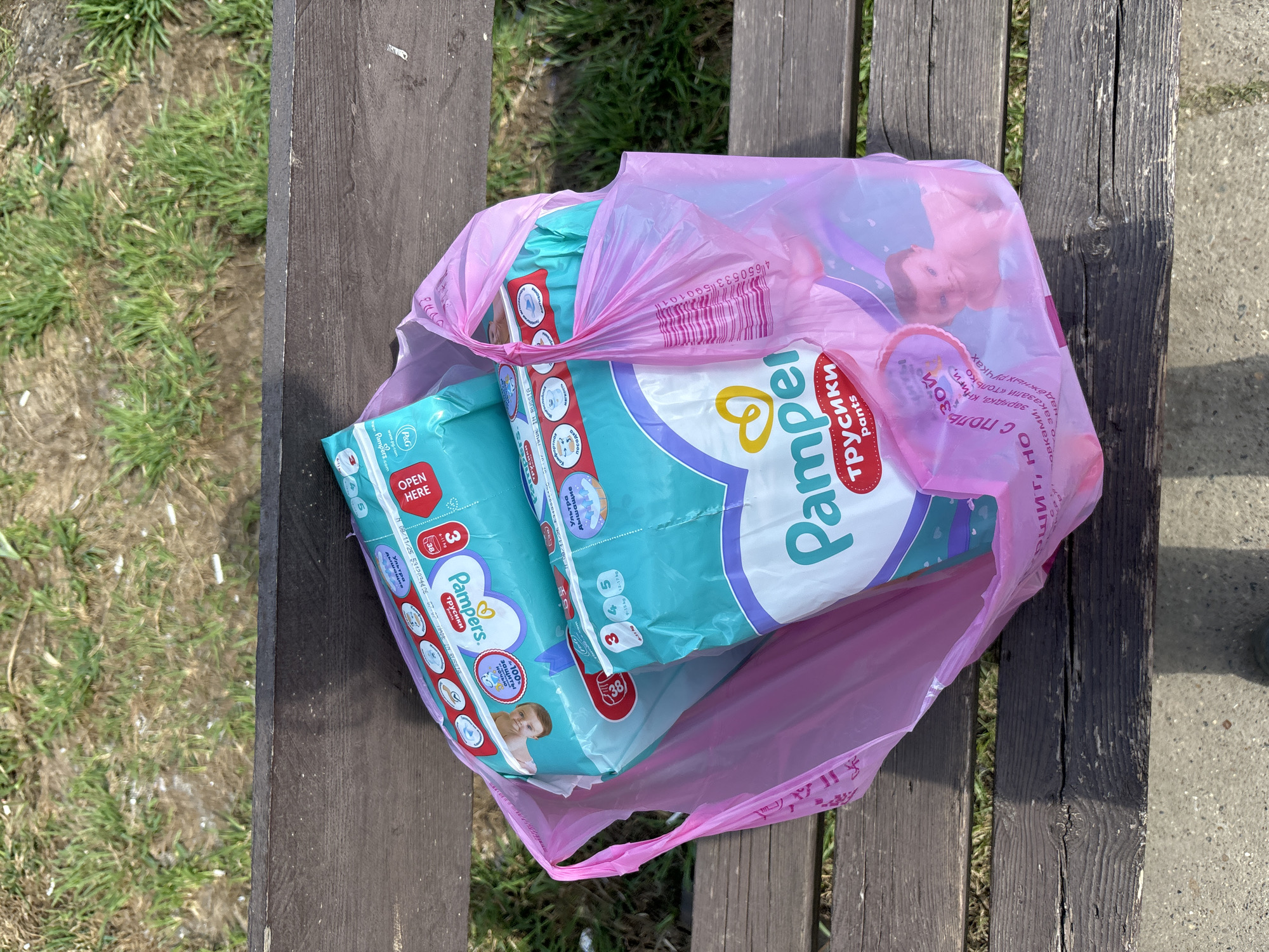 The image depicts a pink plastic bag filled with two packages of Pampers Trusiki baby diapers placed on a wooden bench. The diaper packages are predominantly teal and white with purple accents, showcasing the Pampers logo and branding in Cyrillic script. The pink plastic bag also displays images of a baby and product information.
The bench is weathered, with visible grain and coloration variations in the wood. The setting includes a patch of grass adjacent to the bench, and concrete pavement is visible on the right side of the frame.
The OCR information on the packaging suggests these diapers are manufactured in Russia and contain contact information related to Proctor & Gamble, as well as details on the product such as size, weight range, and certifications of protection. While the OCR provides manufacturer details, there is no clear indication that the image was taken in Markovskiy, Russia.