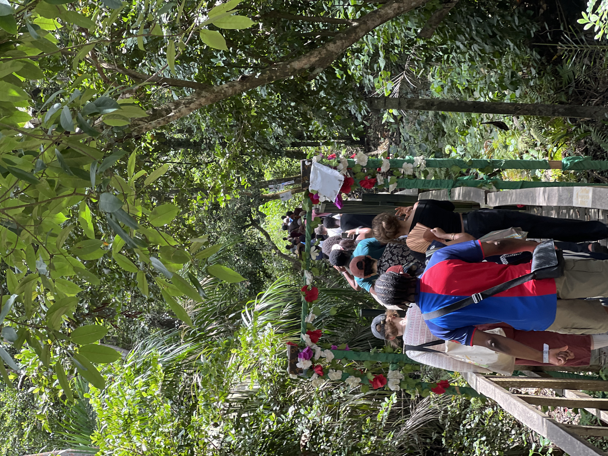 The image shows a group of people on a decorated wooden walkway or bridge in a lush, green environment, possibly a forest or park setting. The walkway is decorated with flowers and green cloth, suggesting a special event or ceremony. The location is identified as Lekki, Nigeria. The people are dressed in a variety of casual clothing and appear to be walking in the same direction. There are trees and vegetation on either side of the walkway, creating a dense, natural setting. Some individuals are carrying bags or fans.