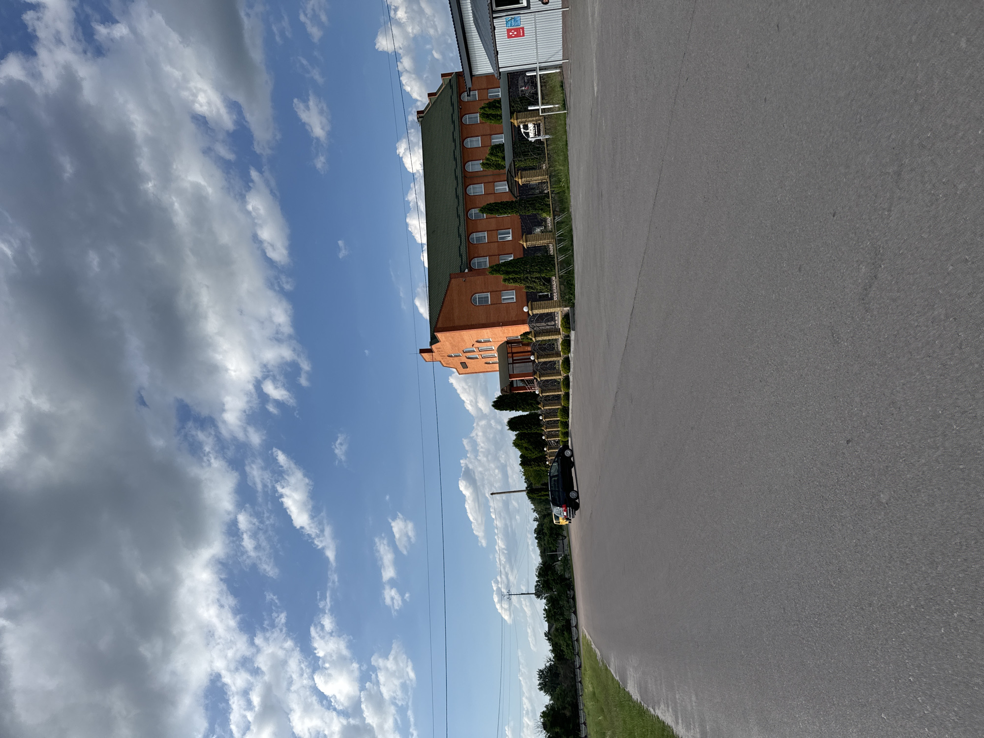 The image depicts an exterior scene in Dovhe, Ukraine, likely during the daytime with fair weather. The sky is filled with a mix of white and grey clouds against a blue backdrop. A two-story building with an orange brick facade and a dark green roof dominates the left side of the frame. The building has numerous windows, some arched. Adjacent to the building, a paved road stretches towards the right side of the image. On the road, a small, dark-colored car with yellow accents is parked. The building is bordered by a low fence and some greenery, including small trees. Power lines are visible in the distance. The overall impression is that of a commercial or residential area with a straightforward, functional aesthetic. No people are visible in the frame.