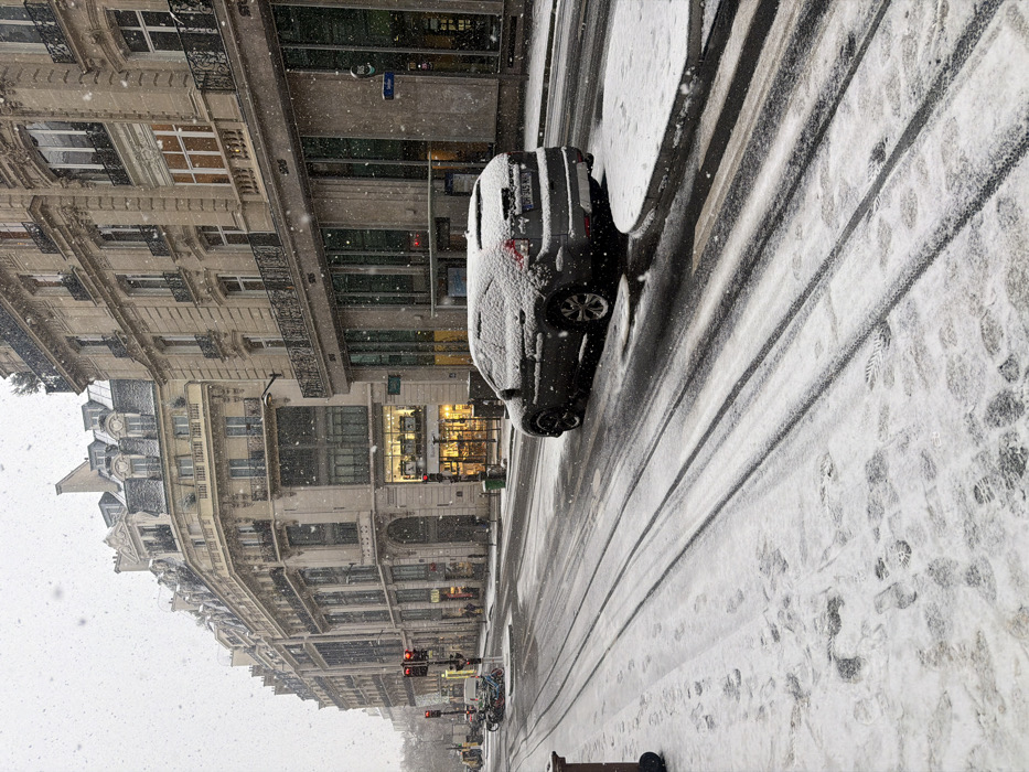 The image depicts an urban street scene in Paris, France, during a snowfall. The location is identified as Quai Anatole France, near the Solférino bus stop and the Ibis hotel, in the 7th arrondissement.
Snowflakes are visibly falling, and a layer of fresh snow covers the street, sidewalks, and parked vehicles. Distinct tire tracks cut through the snow on the roadway, extending from the foreground into the distance. Numerous footprints are present on the snow-covered sidewalk in the foreground and along the left.
On the right side of the street, a dark gray SUV is parked, its roof, hood, and rear covered in snow. Its rear license plate, "DH-345-WW," is visible. A circular, snow-covered concrete barrier or planter is on the sidewalk adjacent to the SUV.
The left side of the street is lined with multi-story, light-colored Haussmannian buildings featuring ornate facades, balconies, and mansard roofs. One building displays a brightly lit storefront with "PAUL" signage, indicating a bakery. On the right, a modern building with large glass panels at ground level features an "Ibis" hotel sign, accompanied by "bus Solferino" text and a directional arrow.
In the distance on the left, a red traffic light is active, and a cluster of motorcycles or scooters are parked near a white van. Several indistinct human figures are visible on the sidewalk in the mid-ground, near the "PAUL" establishment. A green public trash bin is also visible on the sidewalk.