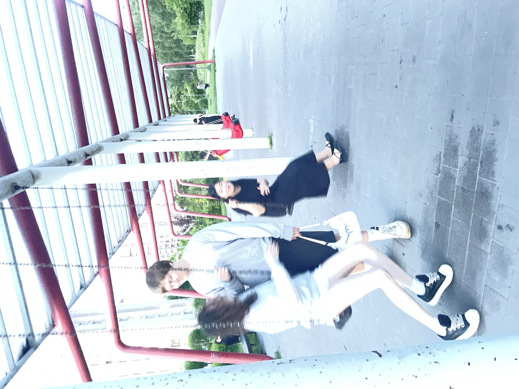 This is a medium shot of a playground in Sinyavino, Russia, with an overhead structure featuring red beams and a white translucent roof. In the foreground, a young woman in a light blue top and white shorts is on a swing, her legs bent. She is blurred due to motion. Beside her, a young man in a grey sweatshirt and black shorts stands holding a white purse. He is looking down and slightly to the side, also slightly blurred. In the mid-ground, another young woman in a black dress and black shoes is on a red swing, her body tilted forward. She is also blurred due to motion. In the background, further down the row of swings, a person in a dark shirt is on a red swing, and another person in a lighter colored shirt is standing near a swing set. The ground is paved with grey interlocking bricks. A line of trees is visible in the distance, with apartment buildings to the left of the frame.