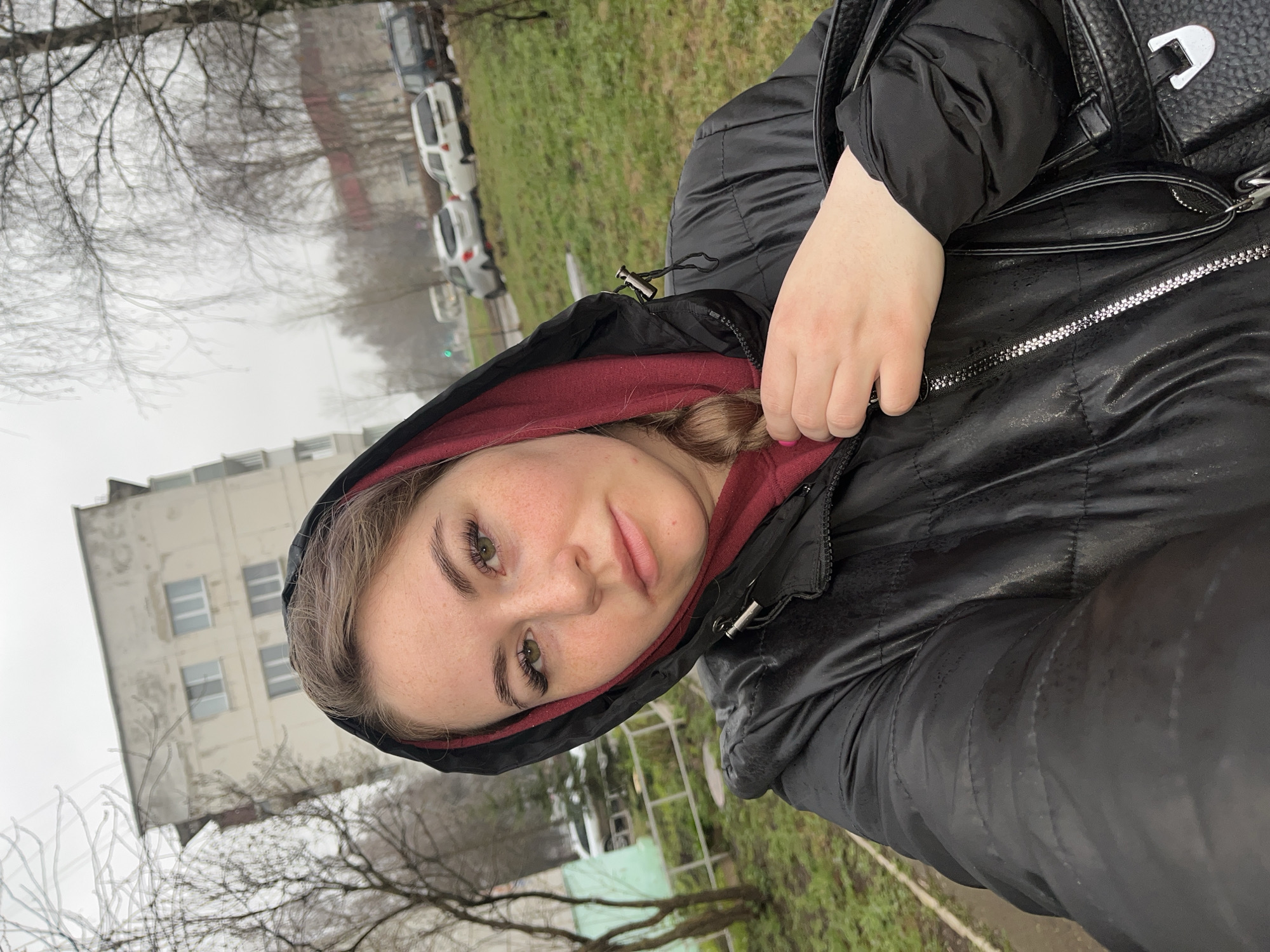 A young woman takes a selfie outdoors in Chaikovsky, Russia, on an overcast day. She is in the foreground, looking directly at the camera with a neutral expression. She has green eyes, prominent eyelashes, and freckles across her nose and cheeks. Her hair, light in color, is partially visible from under the hood. She wears a black quilted puffer jacket with the hood up, revealing a maroon hoodie underneath. Her right hand is visible, resting near the jacket's zipper, with a hint of pink nail polish on one finger. A black shoulder bag with a silver buckle is visible over her right shoulder.
The setting appears to be a residential or urban area, possibly near a park or street. In the background, bare tree branches suggest late autumn, winter, or early spring. A light-colored, multi-story building with multiple windows stands behind the trees. A grassy area and what appears to be a paved road or parking area are also visible, with several parked cars, including white and darker vehicles, lining the street. The sky is uniformly gray, indicating an overcast condition and diffused lighting. No legible text is visible in the image.