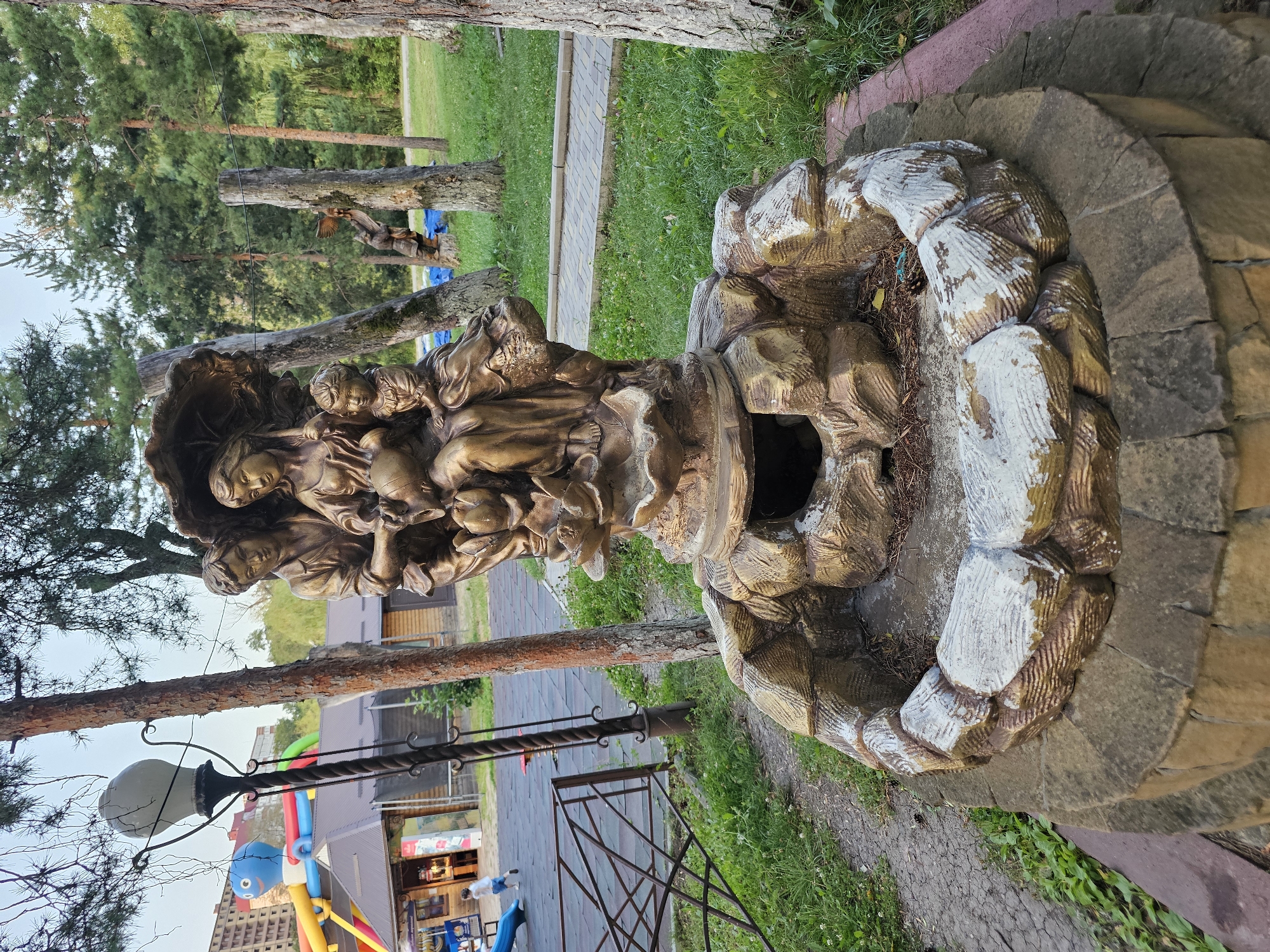 The image shows a bronze fountain sculpture in what appears to be a public park or recreational area. The sculpture depicts a family under an umbrella, with a man and woman holding a baby, and another child at the woman's side. The fountain itself is a circular structure with rough, stone-like ornamentation. In the background, there is a playground with inflatable attractions, a building that appears to be a concession stand or shop, trees, and a paved walkway. A lamppost is visible to the left of the sculpture. The overall setting suggests a family-friendly outdoor space.
The sculpture and fountain are located in Cherkessk, Russia, at the "Zolotoy Kolos" (Golden Ear) park. This park is known for its recreational facilities and attractions for children. The sculpture is a prominent feature within the park, often used as a meeting point or a decorative element.