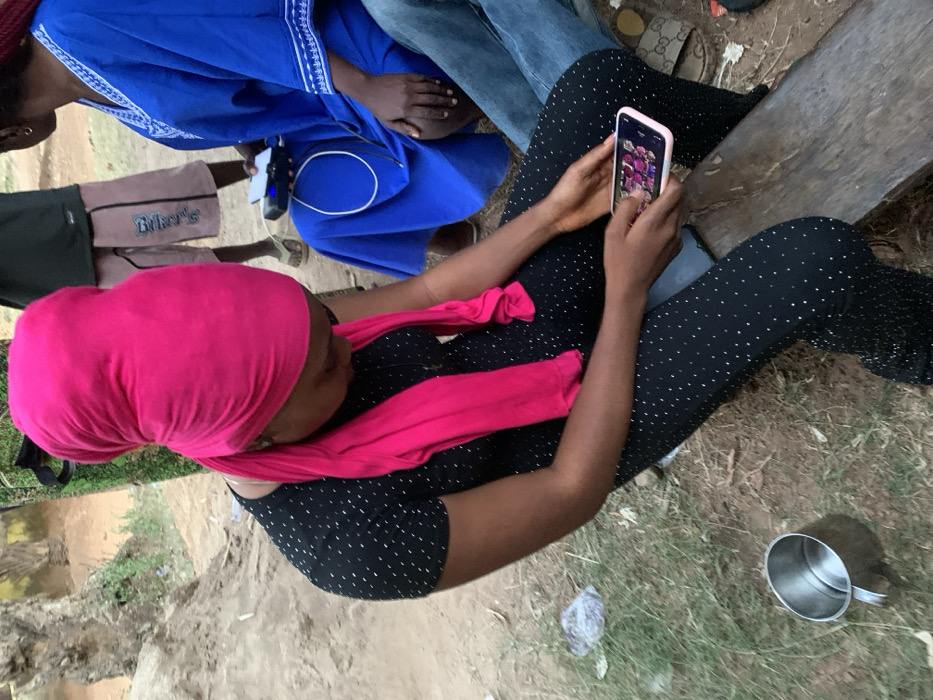 An overhead shot depicts a woman wearing a pink head wrap and a black and white polka dot top and pants. She's sitting outside, holding a phone with a colorful screen. Other people are seated nearby, one wearing blue. A shiny metal mug sits on the ground beside the woman. The ground is dirt and sparse grass.
