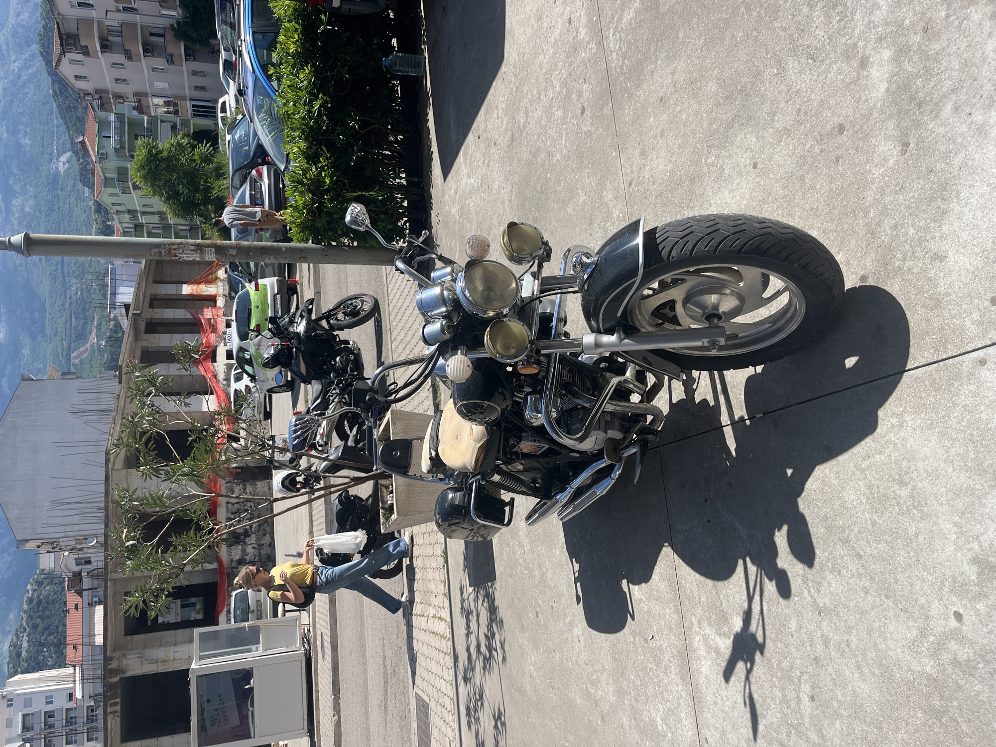 The image depicts a vibrant street scene in Budva, Montenegro, under bright sunshine, indicated by strong shadows. A large, chrome-heavy motorcycle is prominently parked on a concrete sidewalk in the foreground, featuring multiple headlights, a light-colored seat, and a wide front tire with a black fender. Its shadow stretches across the pavement.
To the left of the motorcycle, a woman in a yellow t-shirt and blue jeans is seen standing, holding a white bag. She is positioned near a white kiosk-like structure. Further back, a man wearing a grey shirt and khaki shorts stands near a row of parked cars. One of these vehicles, a green car, is identifiable as a "TAXI" due to visible lettering. Other cars, including a blue and a grey one, are also parked alongside a green bush.
The scene transitions to a midground featuring a multi-story building under construction, marked by rebar and red safety netting, and a small tree. In the background, numerous multi-story residential buildings with balconies are visible, characteristic of a town built into a hillside. Beyond these structures, a vast landscape of green, rolling mountains stretches into the distance under a clear sky.
The only discernible text, apart from "TAXI", appears partially on the white kiosk: "IDEJA PRACA OBAVEZE".