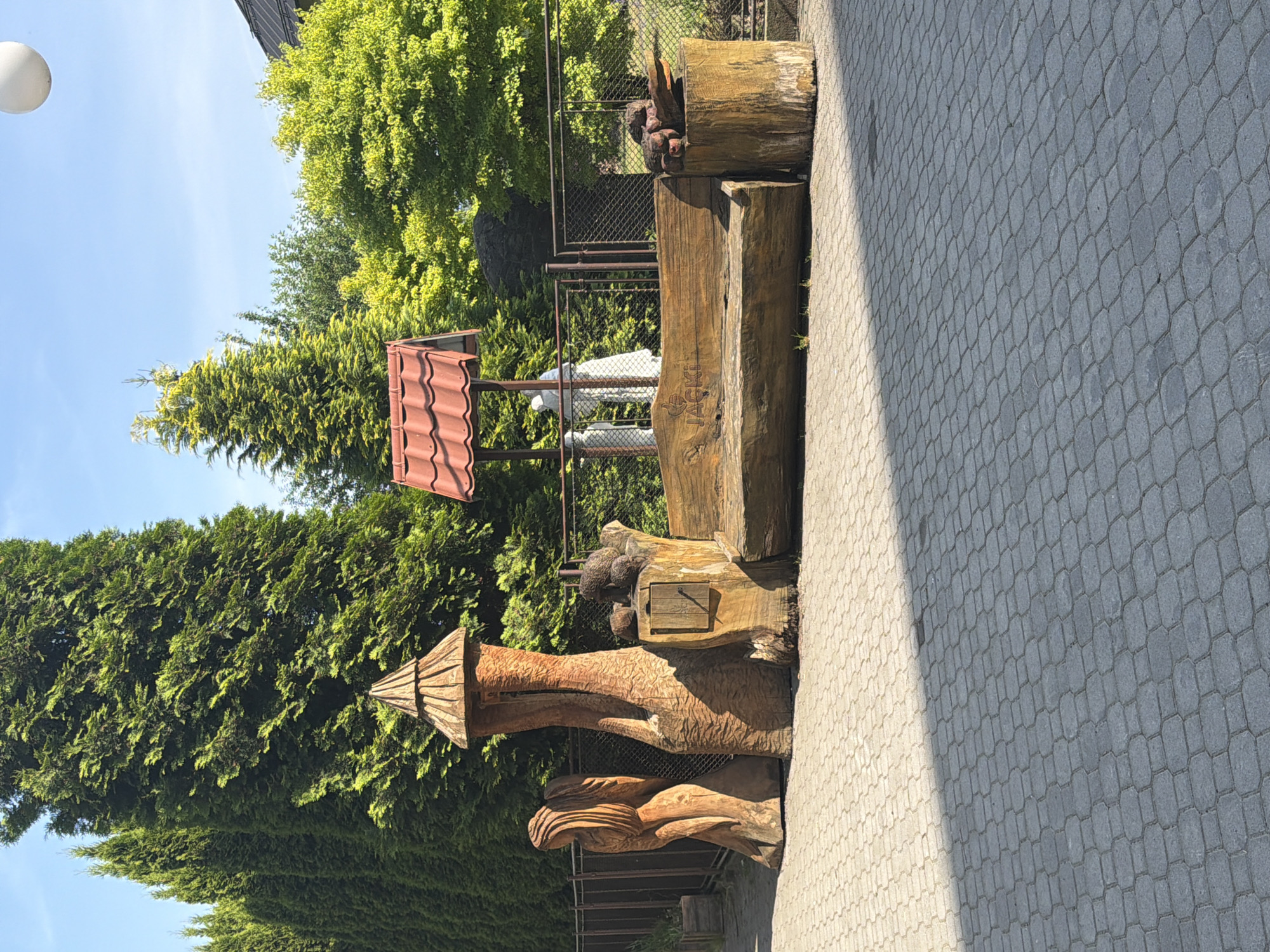 In Jablunkov, Czechia, a paved courtyard features a rustic wooden bench carved into a log. The bench is adorned with carved text reading "JACKI". To the left of the bench, a carved wooden statue of a figure with flowing hair stands next to another carved wooden structure resembling a small tower or stump with a conical roof. To the right of the bench, a cylindrical log serves as a side table, topped with what appears to be a carved animal or wood sculpture. In the background, a fence separates the courtyard from a row of tall, lush green evergreen trees. Behind the fence, a small, red-roofed structure stands, and two figures, possibly statues, are visible beneath it. Sunlight casts shadows across the paved area.