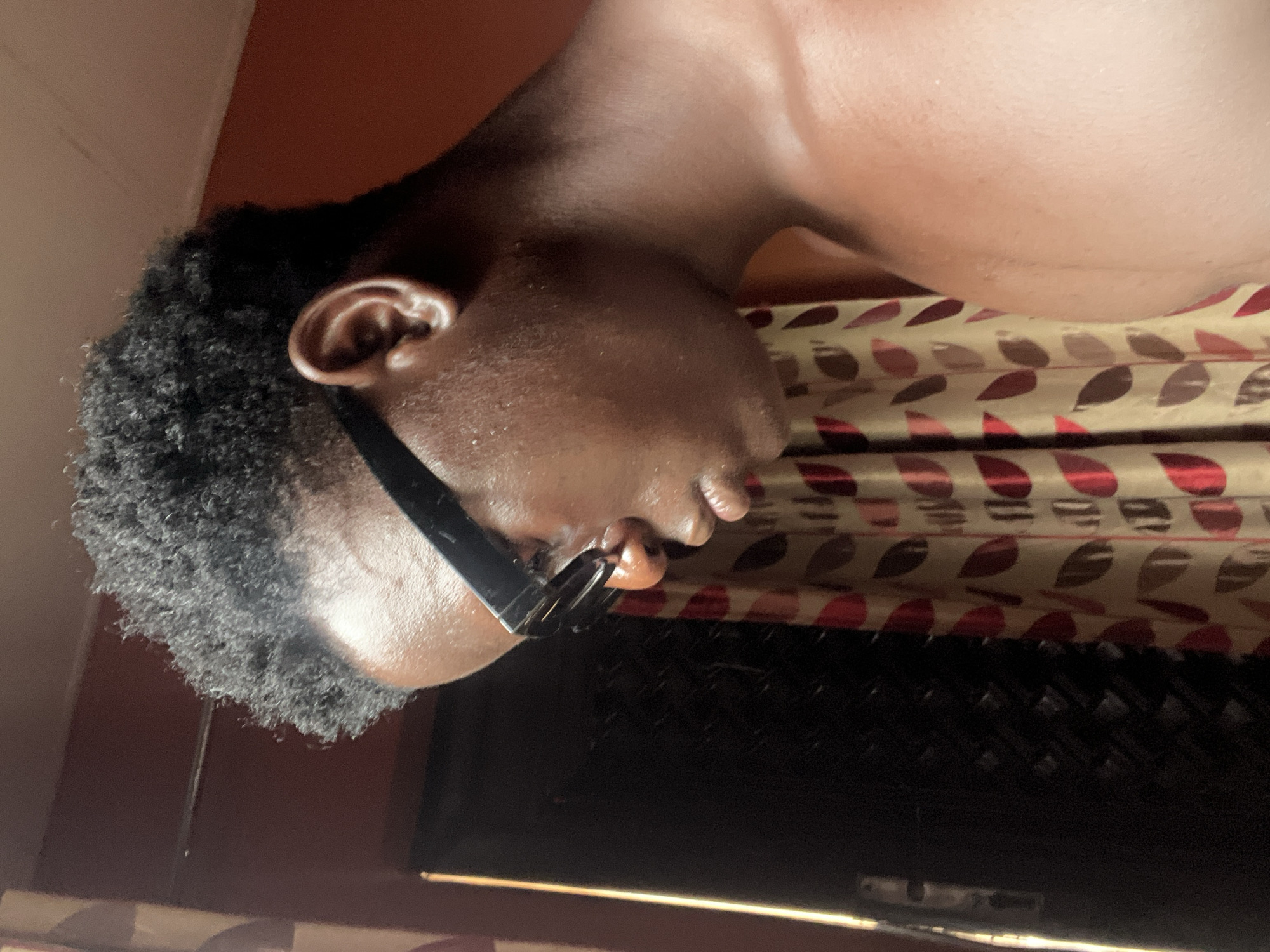A close-up, angled shot depicts a person with dark skin and short, tightly curled black hair. The person is wearing dark-framed glasses. The background includes a window with patterned curtains of gold, red, and brown, and what appears to be the interior wall of a building. There is no information to determine the location of the picture.