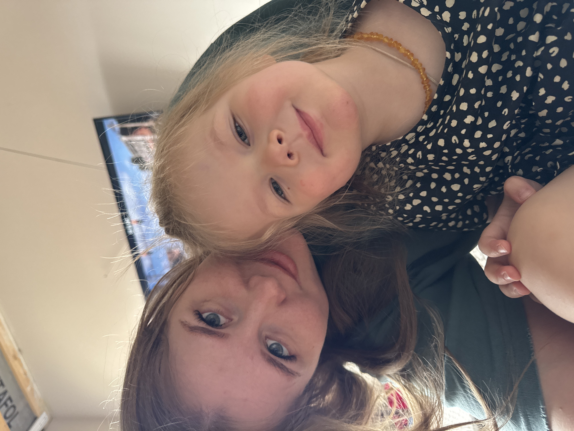 A selfie shows a woman and a young girl. The woman has long, light brown hair and blue eyes. The girl has blonde hair, blue eyes, and is wearing a dark dress with white spots and an amber-colored necklace. They are positioned head-to-head. A television screen is visible in the background. The location is likely indoors. The information about Gornyi (Dvurechenskoe S/P), Russia, is not verifiable from the image.