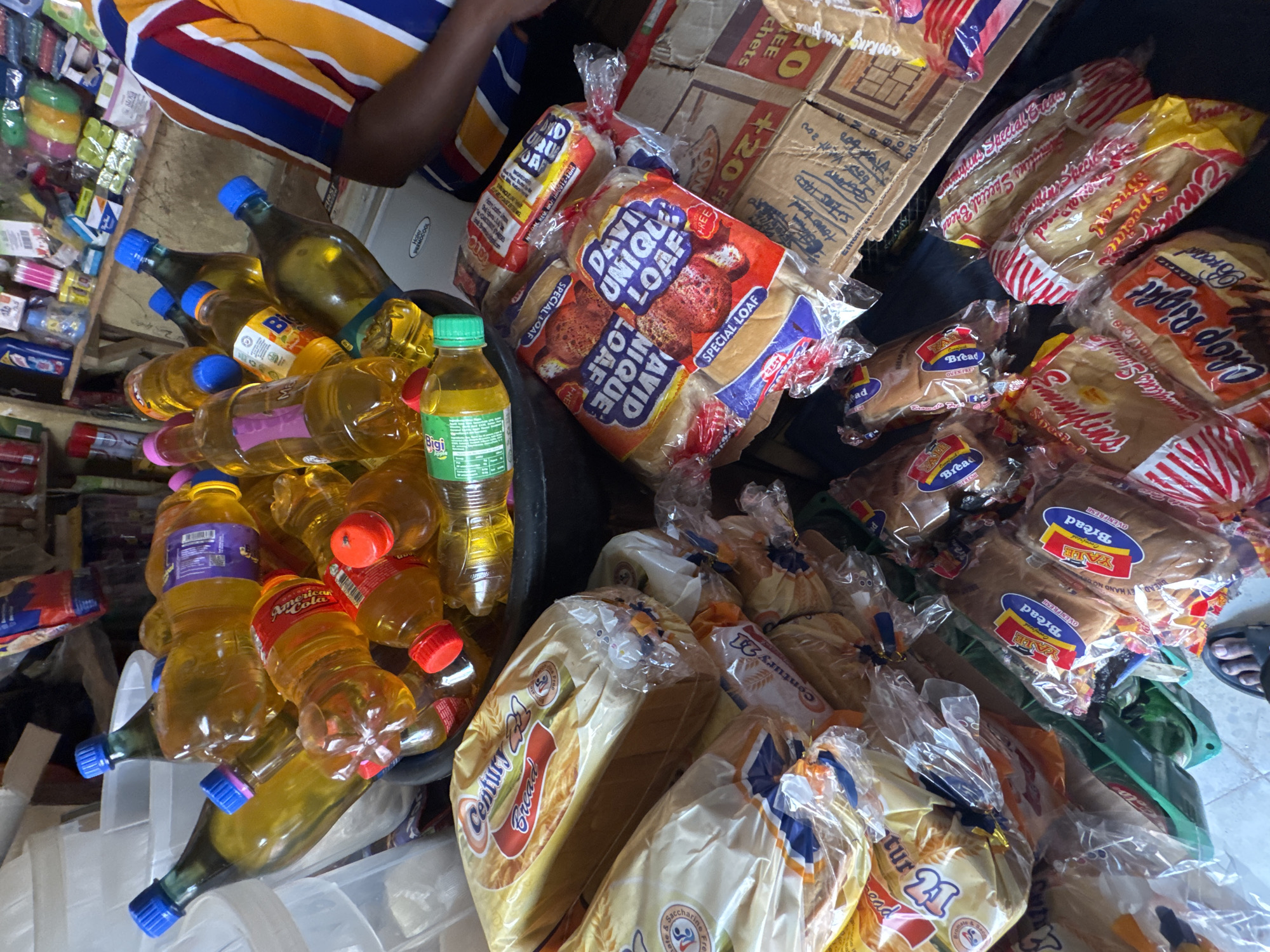 In Ihiagwa, Nigeria, a close-up overhead shot captures a bustling market scene filled with an assortment of food items. The foreground is dominated by stacks of bread loaves, some still in their plastic wrappers, others partially obscured. The bread packaging features various brand names, including "Century 21 Bread," "David Unique Loaf," and "Chop Right Bread." Interspersed among the bread are numerous bottles of cooking oil, their contents a vibrant yellow, with an assortment of colored caps. These bottles are piled high, creating a sense of abundance. In the background, shelves are lined with more products, including detergents and other household items, hinting at the wide variety of goods available in this Nigerian market. A person's arm, wearing a brightly striped shirt, is visible on the right side of the frame, suggesting active participation in the commerce. The lighting is bright, illuminating the scene and highlighting the textures and colors of the products.