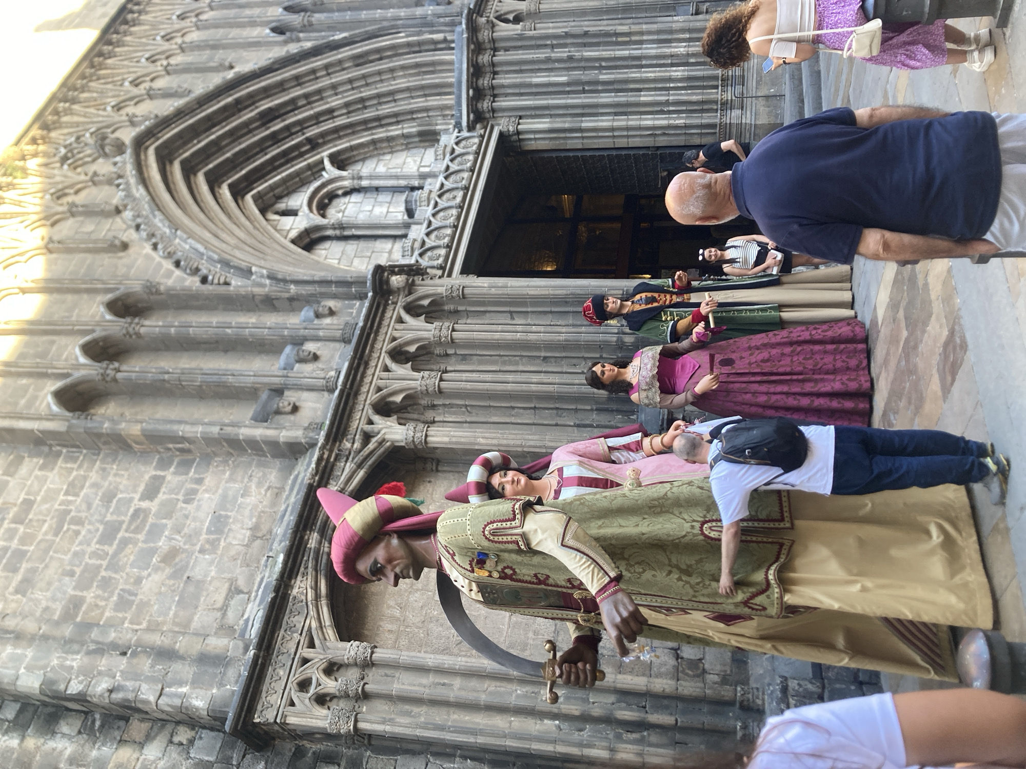 The image captures a vibrant street scene in Barcelona, Spain, featuring several large traditional figures known as "gigantes" (giants) set against the intricate Gothic architecture of a historical building, likely a cathedral or church.
In the foreground, a prominent male gegant, dressed in a green and gold tunic with a red and gold striped turban and holding a curved sword or scimitar, stands tall. Behind him, other gegants are visible, including a female figure in a pink and white striped dress and turban, and another in a maroon and gold dress. These figures are elaborate and represent historical or mythical characters, commonly used in Catalan festivals and processions.
Several people are observing the gigantes. In the middle ground, a bald man in a dark blue polo shirt and striped shorts stands with his back to the viewer, looking towards the figures. Further back, another man with a backpack wears a white t-shirt and blue pants, also observing. On the right, a woman with curly hair, wearing a white top and a purple skirt, is seen looking at her phone. Other individuals are scattered around, casually taking in the scene.
The setting is an impressive stone building with multiple arched doorways and ornate carvings, characteristic of Gothic architecture. The sunlight casts soft shadows, indicating it is daytime, likely late morning or afternoon. The ground is paved with patterned stone blocks. The overall atmosphere suggests a cultural event or display, with tourists and locals gathered to admire the traditional figures. No visible text is clearly discernible in the image.