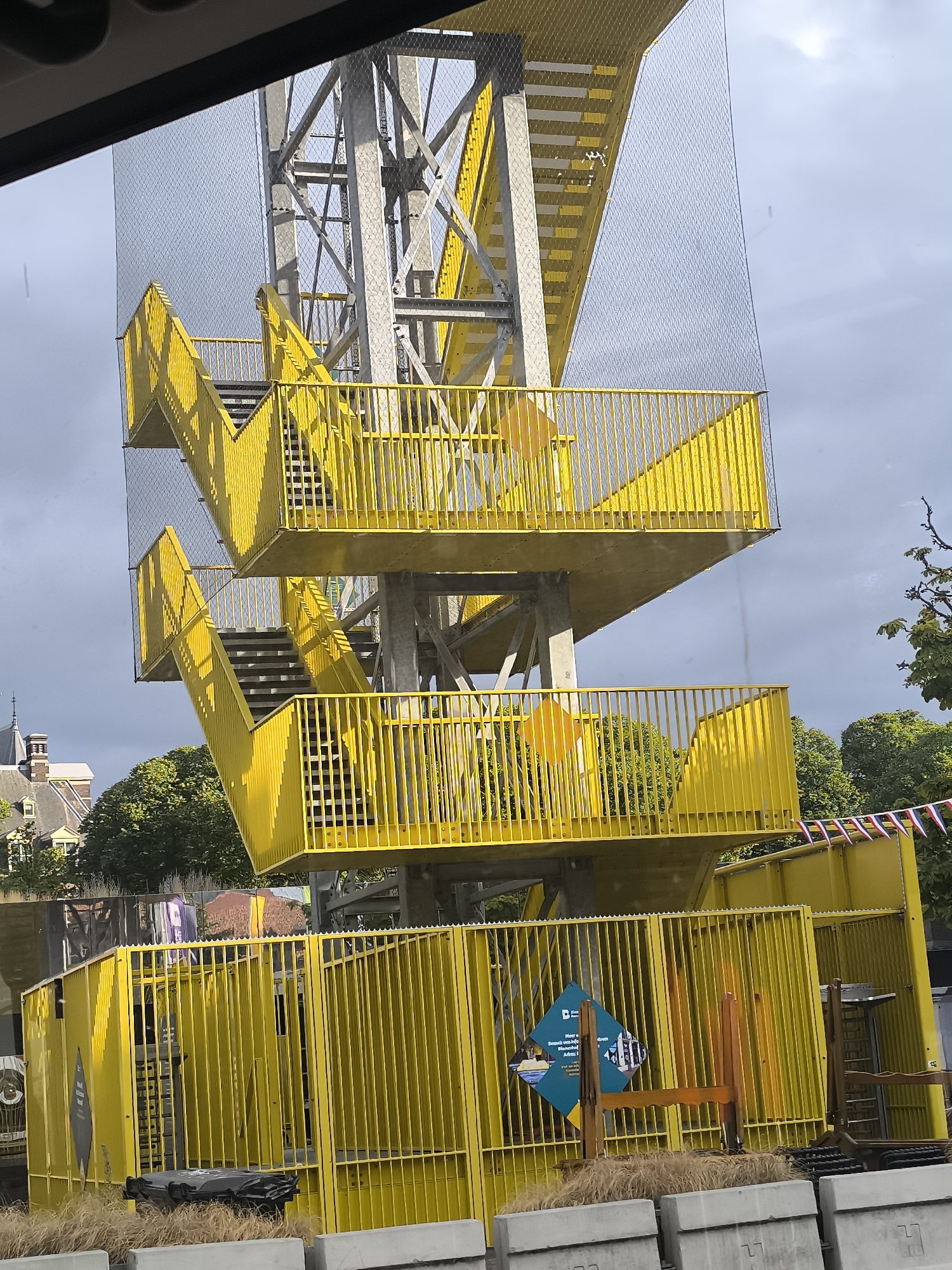 The image shows a tall, bright yellow metal staircase structure with multiple landings and railings, likely part of a playground or observation tower. The structure is supported by a central grey metal frame. A fine mesh netting encloses parts of the upper structure. Below the main staircase, there are yellow fenced-off areas, some containing wooden elements and a sign. In the background, trees and a building with a pitched roof and chimneys are visible. A string of small flags, some appearing to be Dutch, is hung along the top of a yellow structure in the lower right portion of the image. The sky is overcast with patches of sunlight. The scene is located in The Hague, Netherlands, specifically at the Skyview Park.