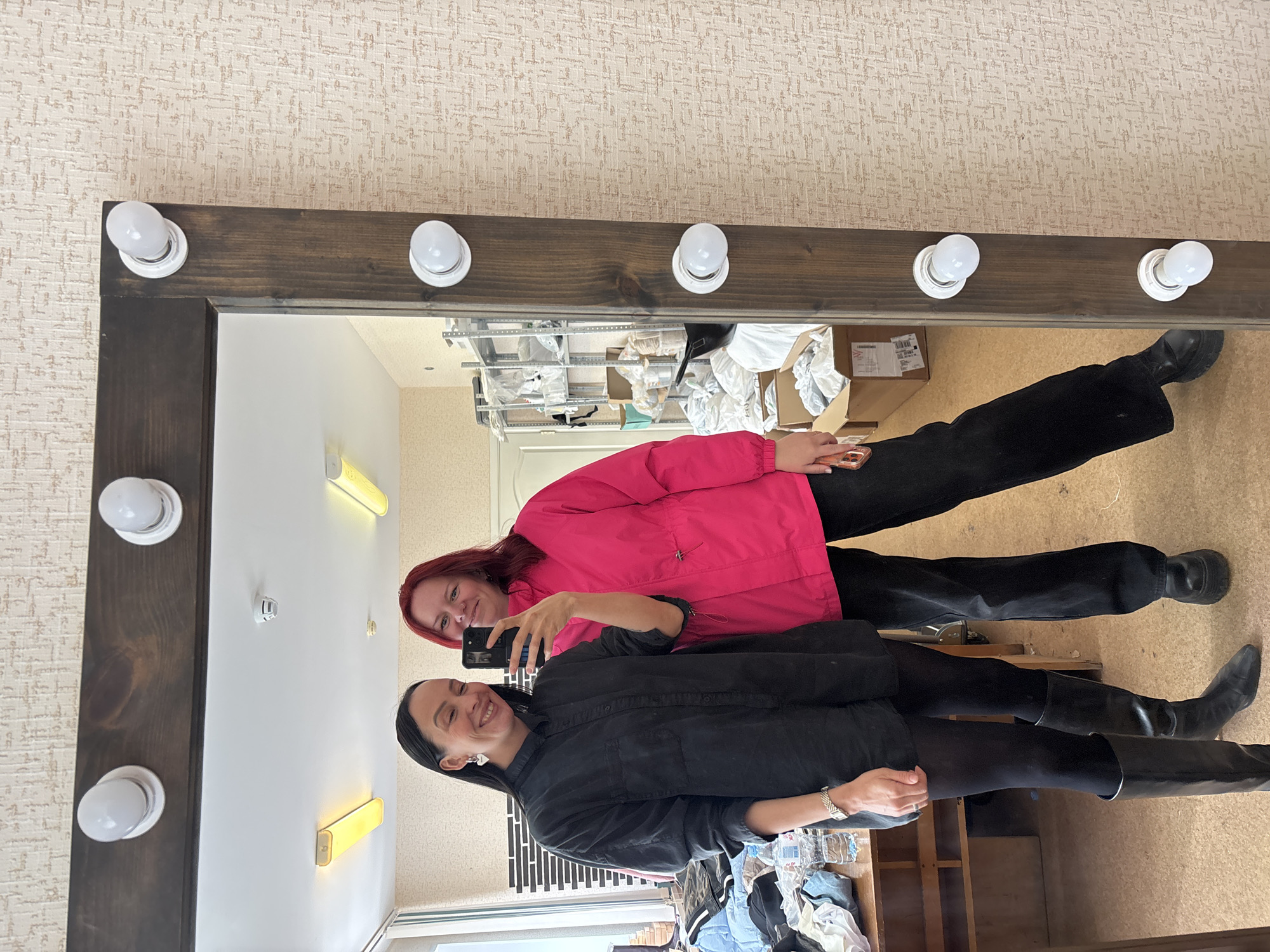 Two women are depicted in a selfie reflection captured in a large, vertically oriented mirror located indoors in Chaikovsky, Russia. The mirror features a dark brown wooden frame with multiple white spherical light bulbs attached uniformly along its perimeter. The woman on the left, with dark hair, wears a black long-sleeved shirt, black tights, black knee-high boots, a silver watch, and heart-shaped earrings. She holds a black smartphone in her right hand, positioned to capture the reflection, and is smiling. The woman on the right, with red hair, wears a bright pink jacket, black wide-leg pants, and black platform shoes, also smiling and looking towards the camera.
The environment visible in the mirror's reflection includes: on the left, a cluttered wooden table with items such as clothing and plastic bottles, adjacent to a window offering a view of an outdoor structure. On the right, beyond the women, metal shelving units filled with various boxed items and white bags are visible, suggesting a storage or workspace. A white door is also partially reflected. The room's walls are covered with light-colored, textured wallpaper. The ceiling is white, featuring a white smoke detector and two rectangular fluorescent light fixtures emitting a yellowish glow. The floor is light brown.