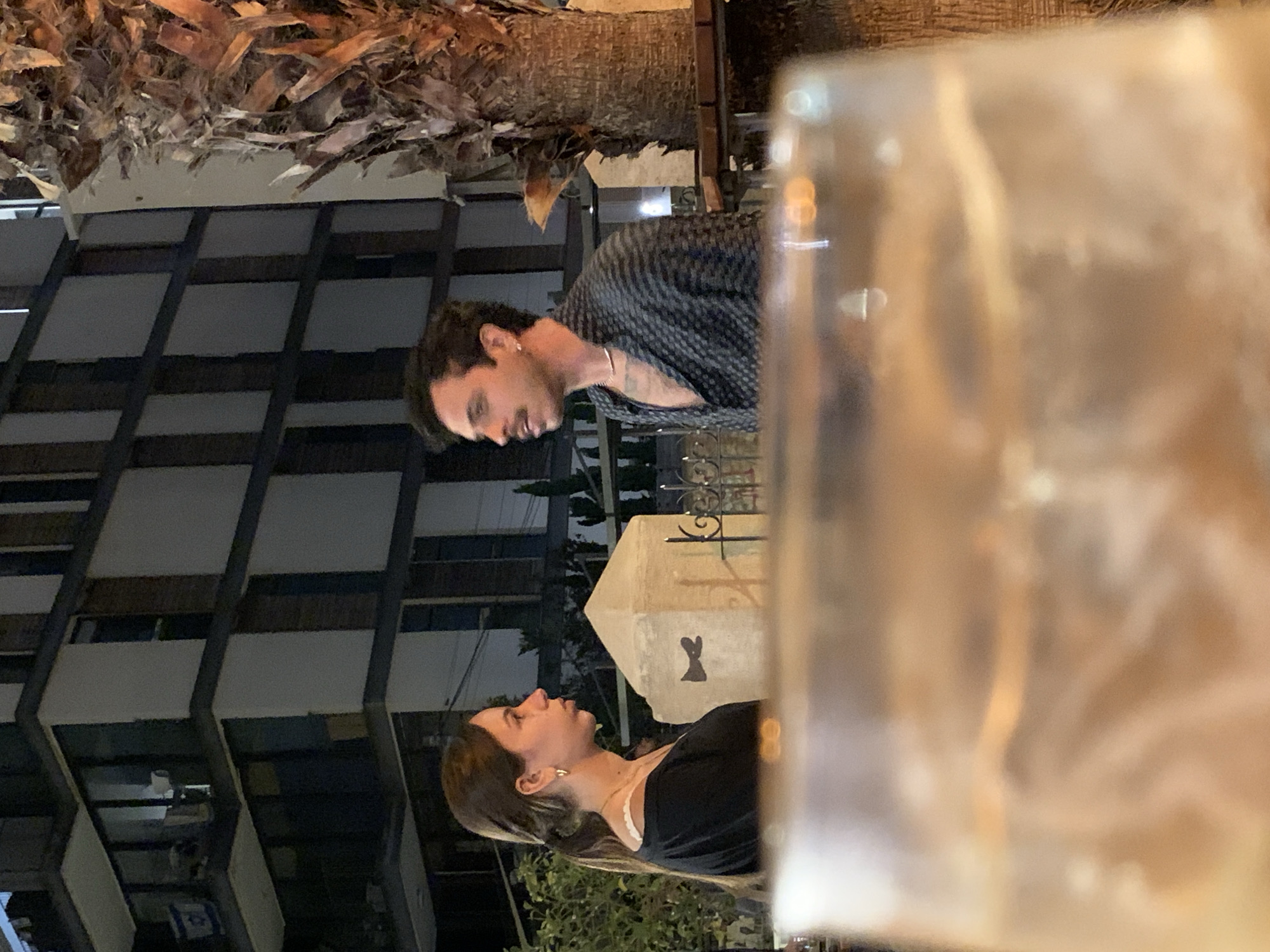 In Tel-Aviv, Israel, a man and a woman are seen in an outdoor setting. The man is wearing a dark patterned shirt and faces left, while the woman, dressed in black, looks towards the right side of the frame. Both are close to each other. The backdrop shows a building with multiple windows, and a palm tree is partially visible above. A street lamp with a rabbit image on it is also in view. A blurry object occupies the foreground, partially obscuring the scene.