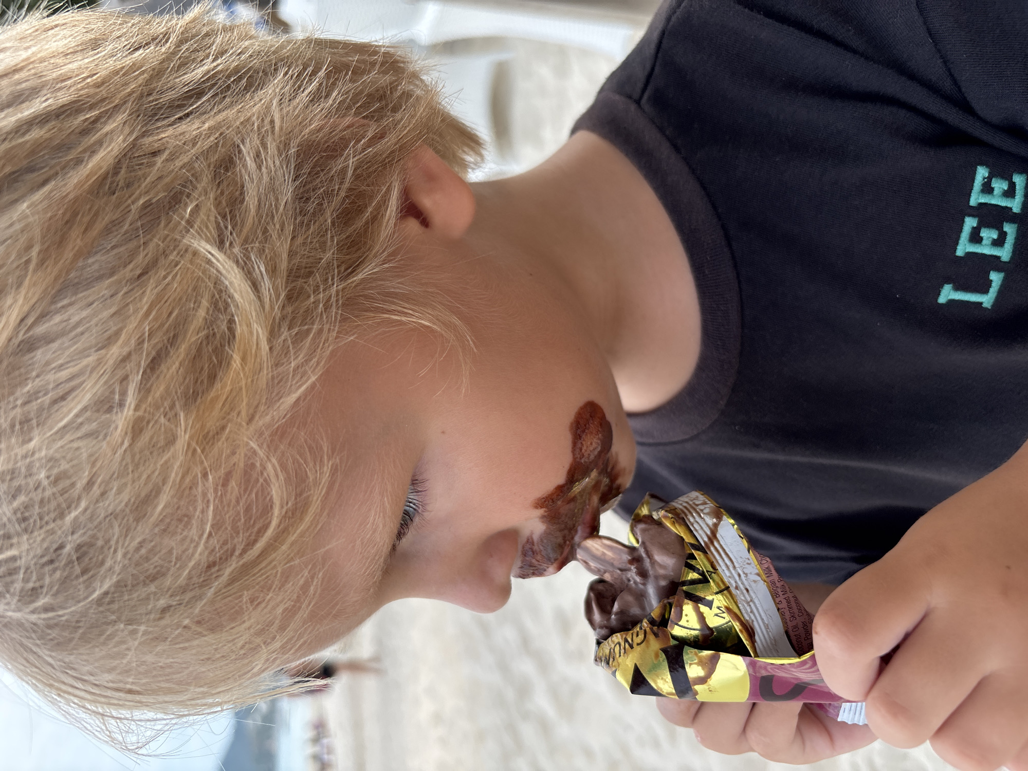 A close-up shot depicts a young boy with blond hair, engrossed in eating a chocolate bar, evidenced by the chocolate smeared on his face. He is wearing a dark-colored T-shirt with the word "LEE" vertically printed on it in a light color. He is holding the partially opened wrapper of the chocolate bar. The background appears to be outdoors, possibly a beach or a similar sandy environment. The location is near Baan Chaweng, Thailand.