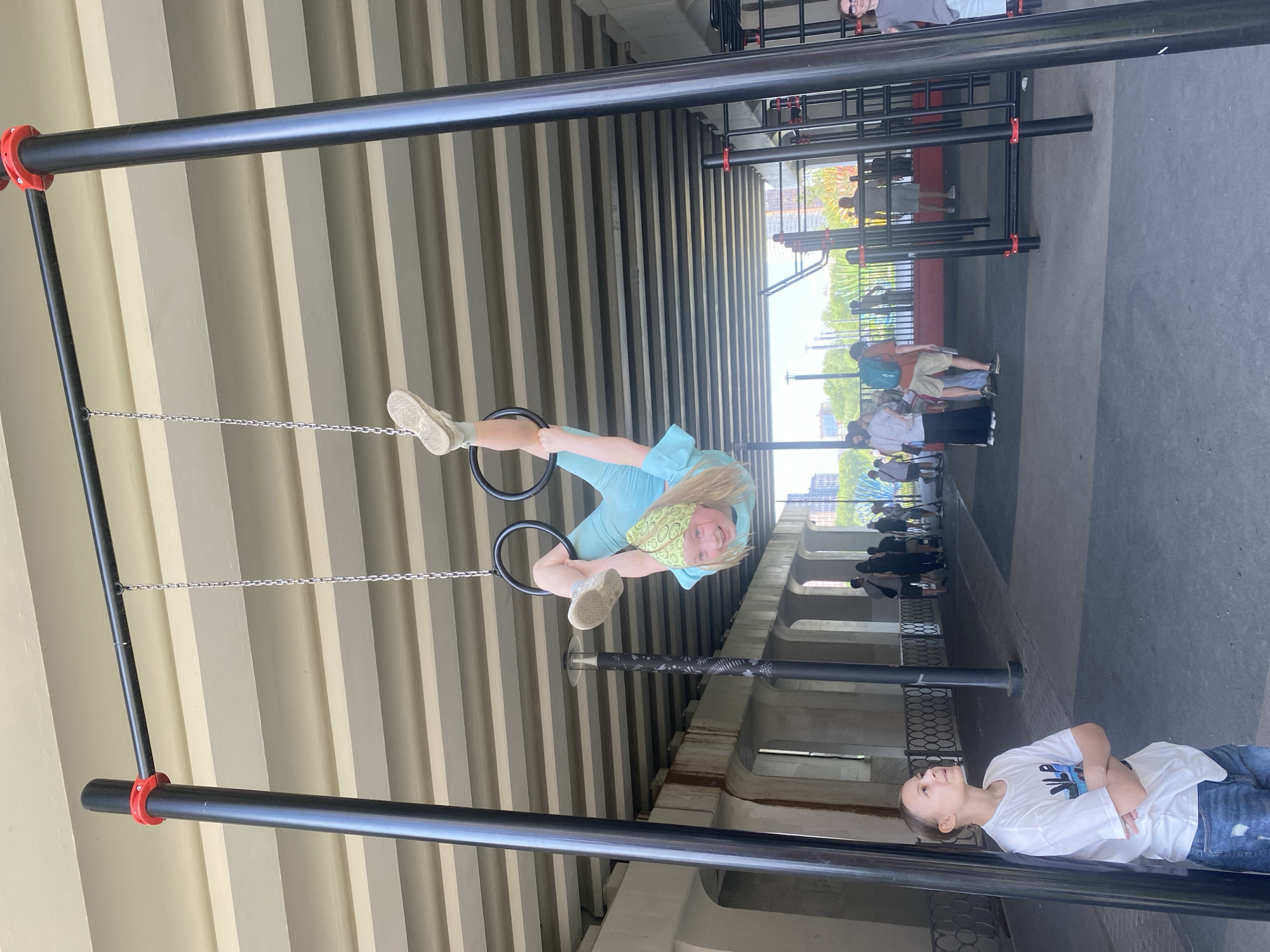 A young girl in a light blue outfit and a patterned head covering is hanging upside down from gymnastic rings suspended from a black metal bar. She appears to be smiling and looking at the camera. Another person, wearing a white shirt and jeans, stands on the ground with their arms crossed, seemingly observing the girl. They are in an open-air exercise area situated under a bridge, identifiable by the repetitive concrete support beams overhead.
In the background, a crowd of people is visible, walking along a riverside promenade. The cityscape of Yekaterinburg, Russia, can be inferred from the architectural style and riverside location, placing this scene near the Iset River, a common recreational area within the city. The photograph appears to have been taken during the daytime under bright, natural lighting.