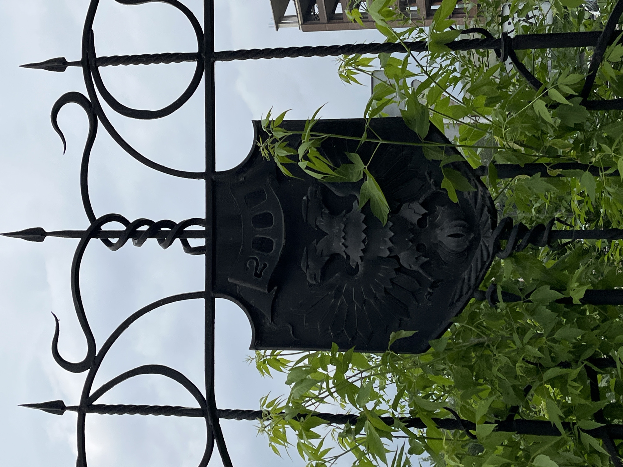 The image shows a black, ornate metal gate with a central shield bearing a double-headed eagle design. Above the eagle, the year "2000" is inscribed. The gate is decorated with twisted metalwork and sharp finials. Lush green foliage, likely climbing vines, is growing around and through the gate, partially obscuring the central emblem. In the background, a multi-story building under construction is visible, with scaffolding and unfinished concrete elements. The sky is overcast. This gate is located at the entrance to the National Centre for contemporary arts (NCCA) in Krasnoyarsk, Russia.
