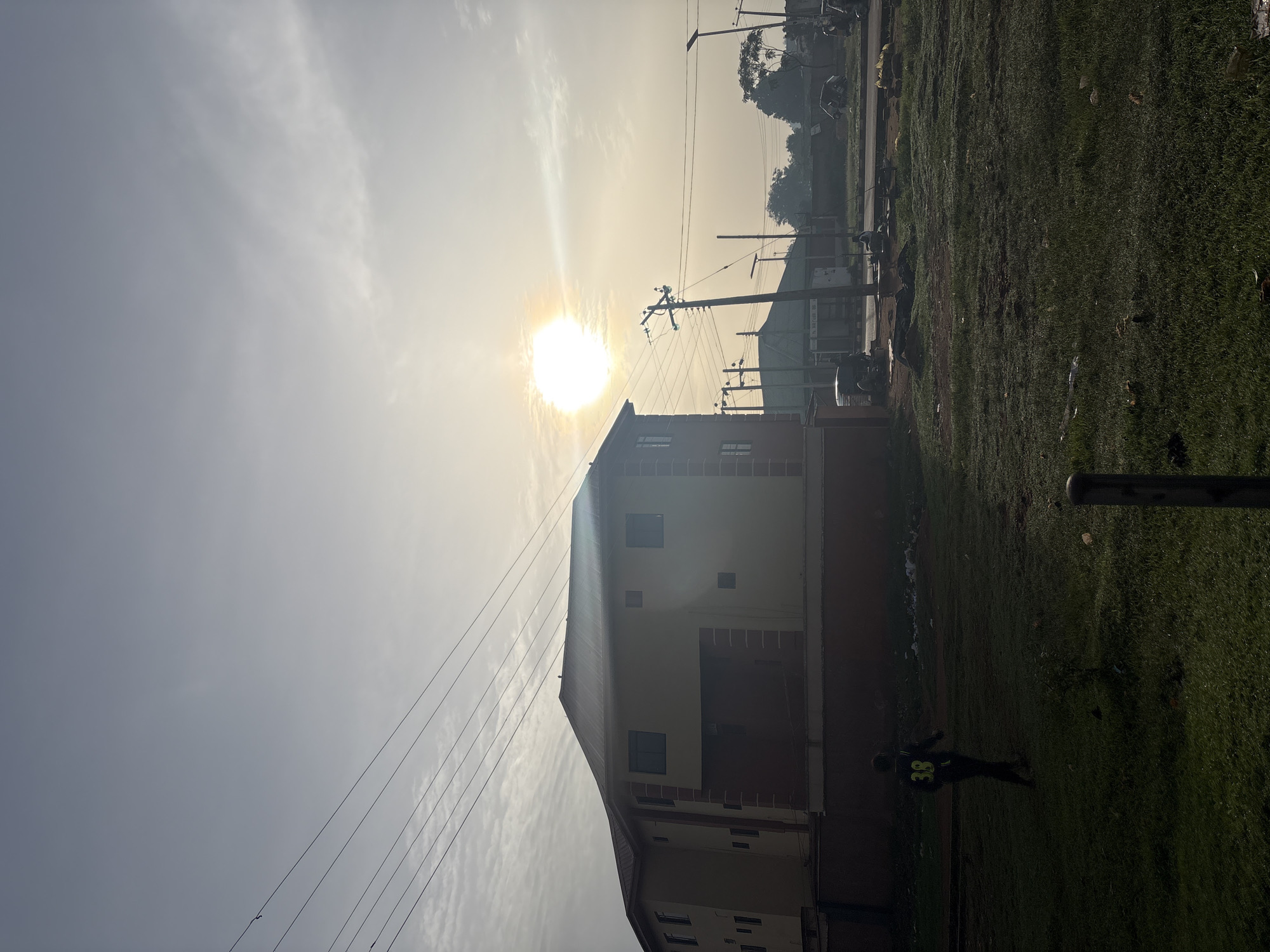 The image depicts an outdoor scene, possibly in Bukuru, Nigeria, characterized by a bright sun in a hazy sky. In the foreground, a person wearing a dark shirt with the number "38" in yellow is walking across a grassy area. To the left, a multi-story building with a reddish-brown foundation and yellow walls stands prominently. Power lines crisscross the sky above the building and extend across the scene. In the background, there are more buildings, streetlights, power poles, and what appear to be several vehicles, including motorbikes and a tricycle-like vehicle. The ground in the foreground is covered in green grass, with some scattered debris. The overall impression is of a moderately developed urban or semi-urban environment during daylight hours, possibly early morning or late afternoon given the sun's low angle and the haze.