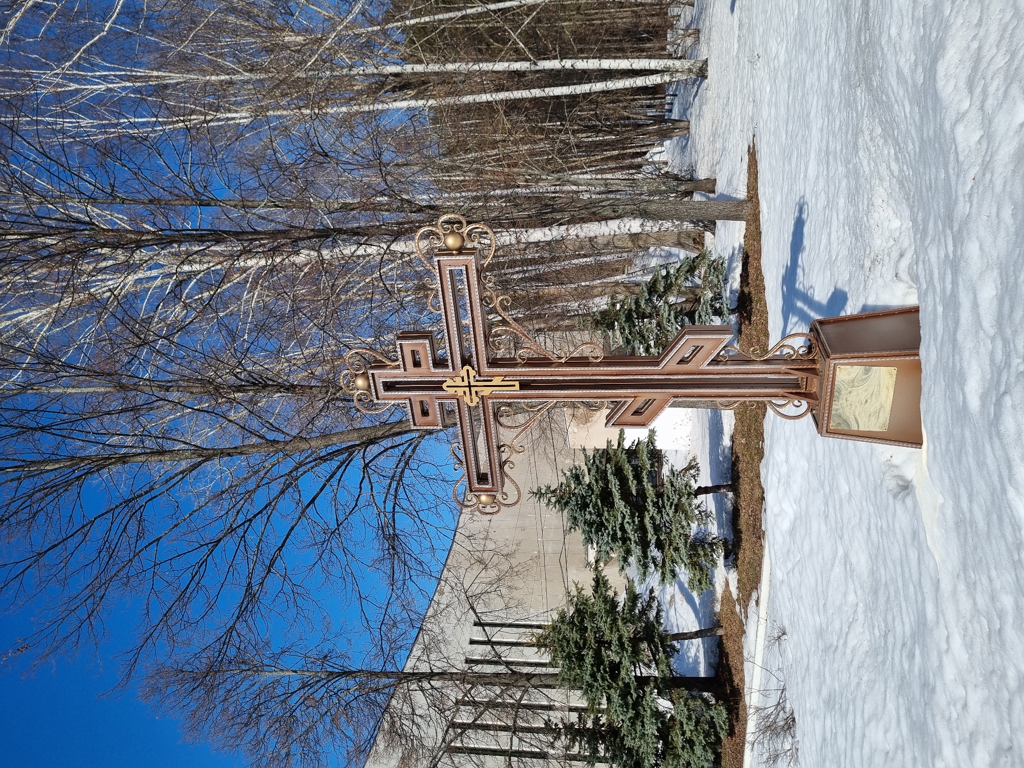 The image depicts an outdoor winter or early spring scene in Chaikovsky, Russia, specifically in the vicinity of the Chaikovsky Art Gallery at Sovetskaya Ulitsa 27. A large, ornate Orthodox cross, constructed of bronze-colored metal with gold-colored accents and decorative scrollwork, stands centrally in a snow-covered landscape. The cross features a stepped upper bar and a diagonal lower bar, resting on a matching rectangular base partially covered by snow.
The ground is predominantly covered in white, textured snow, showing undulations and some areas of exposed brown earth, particularly beneath trees. To the left of the cross, several small, green coniferous fir trees are visible. The background features a dense stand of bare deciduous trees, including numerous birches with distinct white bark, extending from the right of the frame. In the upper left background, a large, light-colored building with a facade characterized by vertical concrete fins is visible; this building is identified as the Chaikovsky Art Gallery. The sky is clear and deep blue. The scene is illuminated by bright sunlight from the left, casting visible shadows on the snow. No individuals, animals, or active events are present.