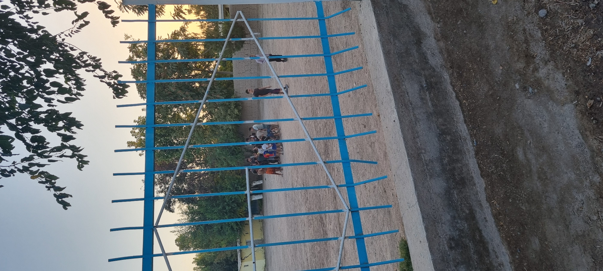The image shows an outdoor scene with a group of young people gathered on a dirt field. The foreground is dominated by a blue metal fence. Beyond the fence, a group of about ten to twelve individuals, predominantly young males, are standing in a cluster. Some appear to be engaged in conversation, while others are looking in different directions. One individual is leaning against a bicycle. In the background, there are trees and a small, light-colored building with a brown roof. The sky is a pale blue with hints of yellow, suggesting it might be late afternoon or early evening. The ground is a mixture of dirt and scattered debris. The overall impression is of a casual gathering in a somewhat unkempt outdoor space, possibly a schoolyard or a local park in the city of Khavaskor, Tajikistan.