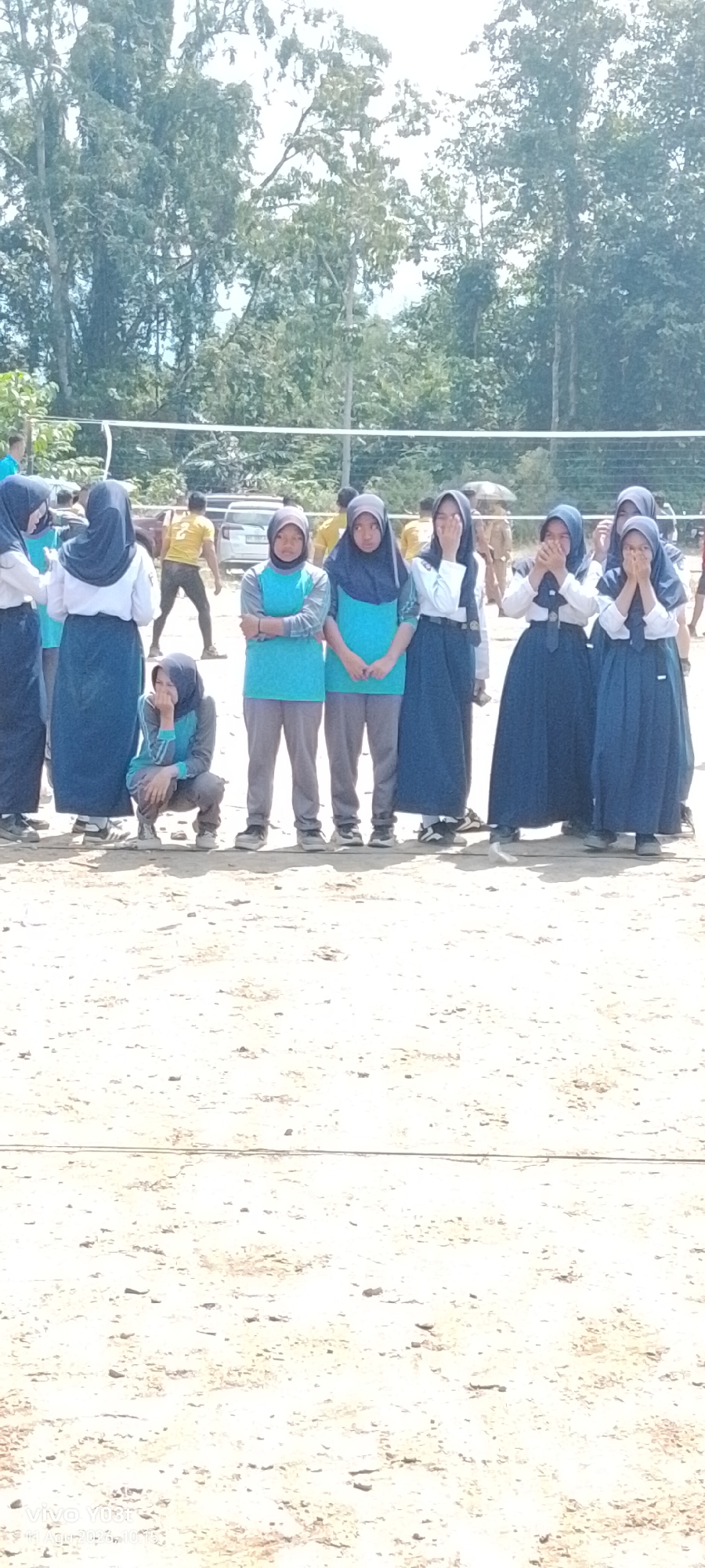In Tirawuta, Indonesia, a group of young women, presumably students, stand in a line outdoors on a sandy or dirt surface, with a volleyball net in the background. They are wearing school uniforms, consisting of long skirts and headscarves. Several of the women have their hands covering their faces, suggesting shyness, laughter, or a gesture of modesty. One individual is crouched in the foreground, also with a hand near their face. Trees and foliage are visible behind the volleyball net, and there are some vehicles and other people in the distance. The overall scene appears to be a casual outdoor gathering or event.