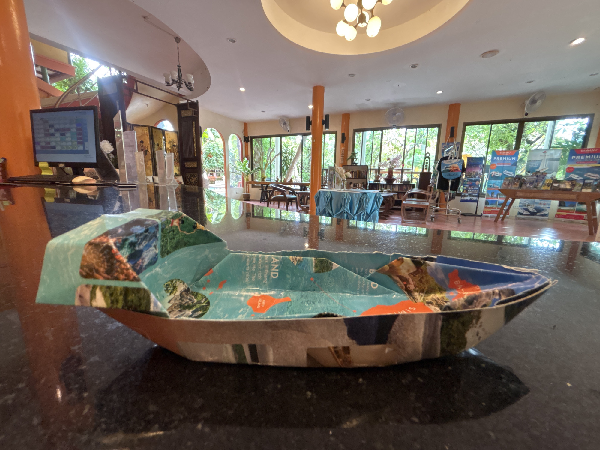 The image captures a close-up view of a handmade paper boat resting on a dark, reflective surface, likely a countertop. The boat appears to be constructed from a colorful material adorned with images of natural landscapes, possibly a map or brochure. In the background, the scene unfolds within what appears to be a reception area or lobby of a resort or hotel in Chalong, Thailand.
The environment suggests a tropical setting, with large windows revealing lush greenery outside. The interior is characterized by warm, orange-toned pillars, a white, domed ceiling with modern lighting fixtures, and a chandelier. The reception desk area features a computer monitor displaying a grid of colored squares, and several clear display stands. Further into the room, tables and chairs are set up, with one table draped in a turquoise cloth. Several promotional posters, possibly advertising tours or packages, are visible on stands in the middle ground. The overall atmosphere is bright and airy, with natural light filtering in from the windows. No people are clearly visible in the image.