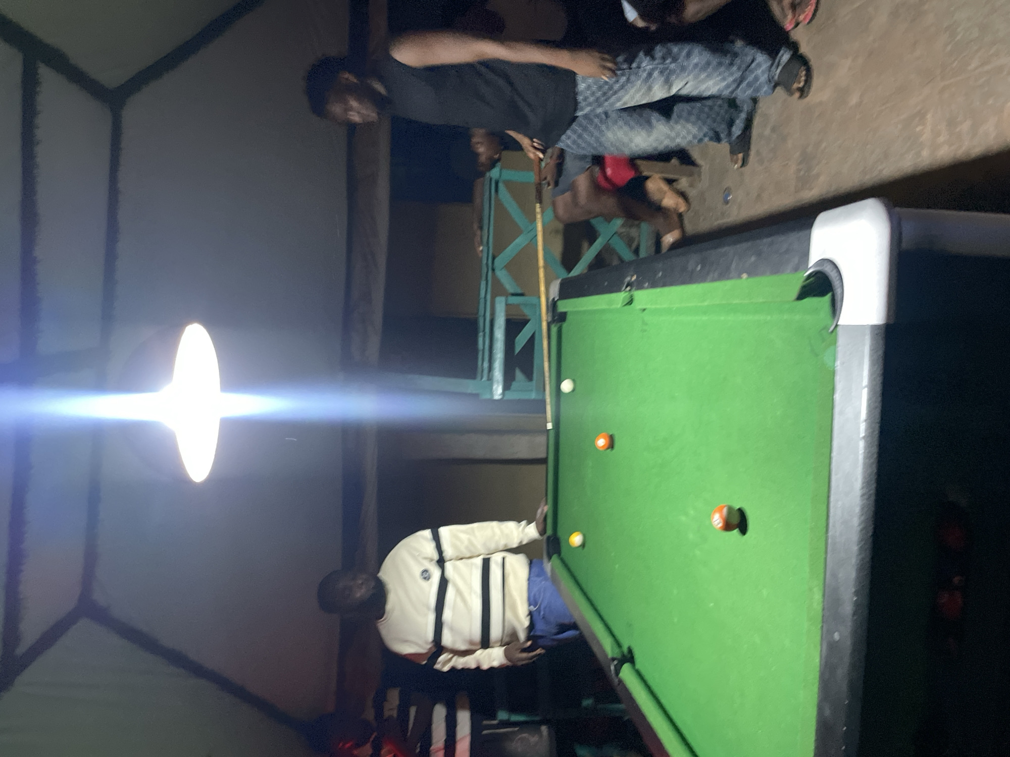 A nighttime scene inside a tent structure in Lokoja, Nigeria, features a billiard table prominently. Two men are present: one wearing a black shirt and jeans stands near the head of the table with a pool cue, while the other, wearing a white and black striped sweater and blue pants, stands near the side of the table. The billiard table is green and contains four balls: one white, one yellow, and two orange. Overhead, a bright light illuminates the scene, casting a glare. A dog is also present in the background. The background includes a partially visible structure of green vertical slats.