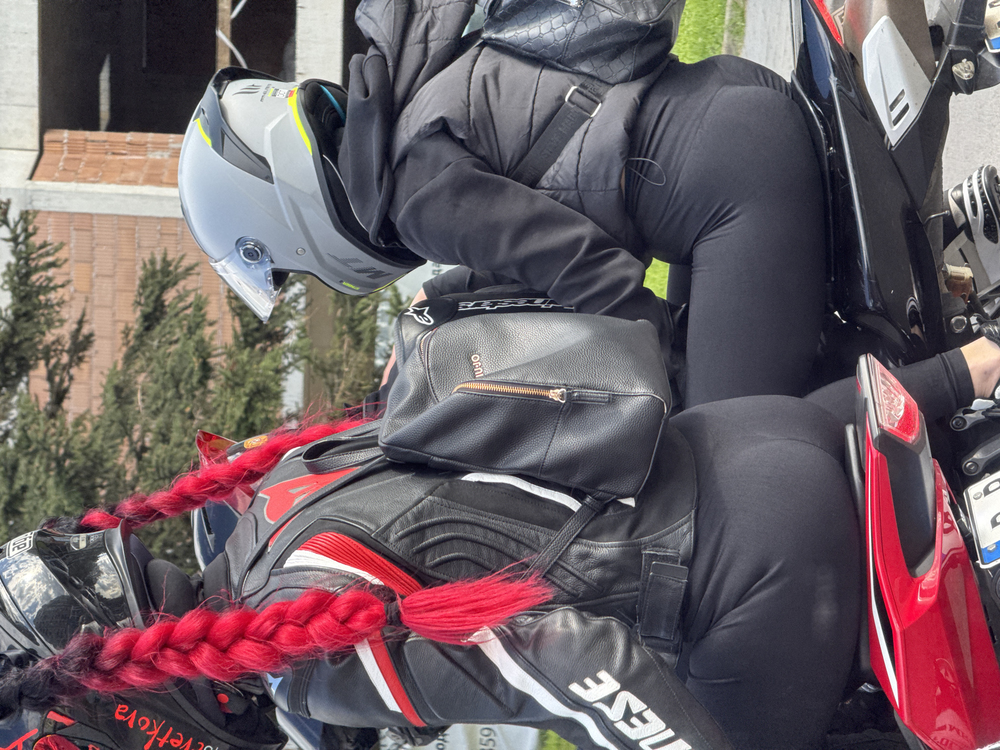 A motorcyclist and a pillion rider are astride a motorcycle. The pillion rider is wearing black leggings and a black padded jacket, a grey helmet, and a small black shoulder bag. The motorcyclist is wearing a black and white leather jacket with red trim, black leggings, a black helmet, and red braided hair extensions. The motorcycle has a red tail light. The background includes trees and a building under construction. The location is likely Varna, Bulgaria, based on a partial license plate visible on the motorcycle.