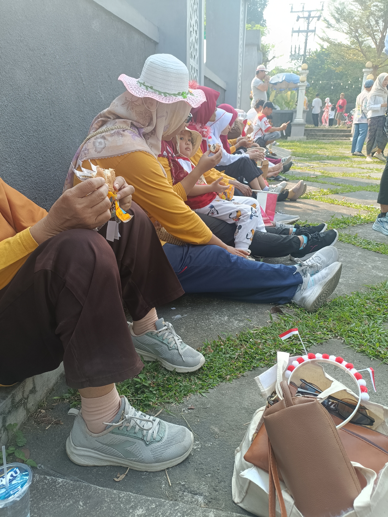 In Sleman, Indonesia, a group of people are sitting on the ground near a building with a textured gray wall and an ornate white structure. In the foreground, a person in brown pants and a yellow long-sleeved shirt is holding a piece of food. Next to them, another person in blue pants and grey sneakers is seated. A bag containing a brown wallet, sunglasses, and small Indonesian flags is placed on the ground. In the background, more people are visible, some standing and some sitting, in a grassy area with trees and a utility pole. The atmosphere suggests an outdoor gathering or event.