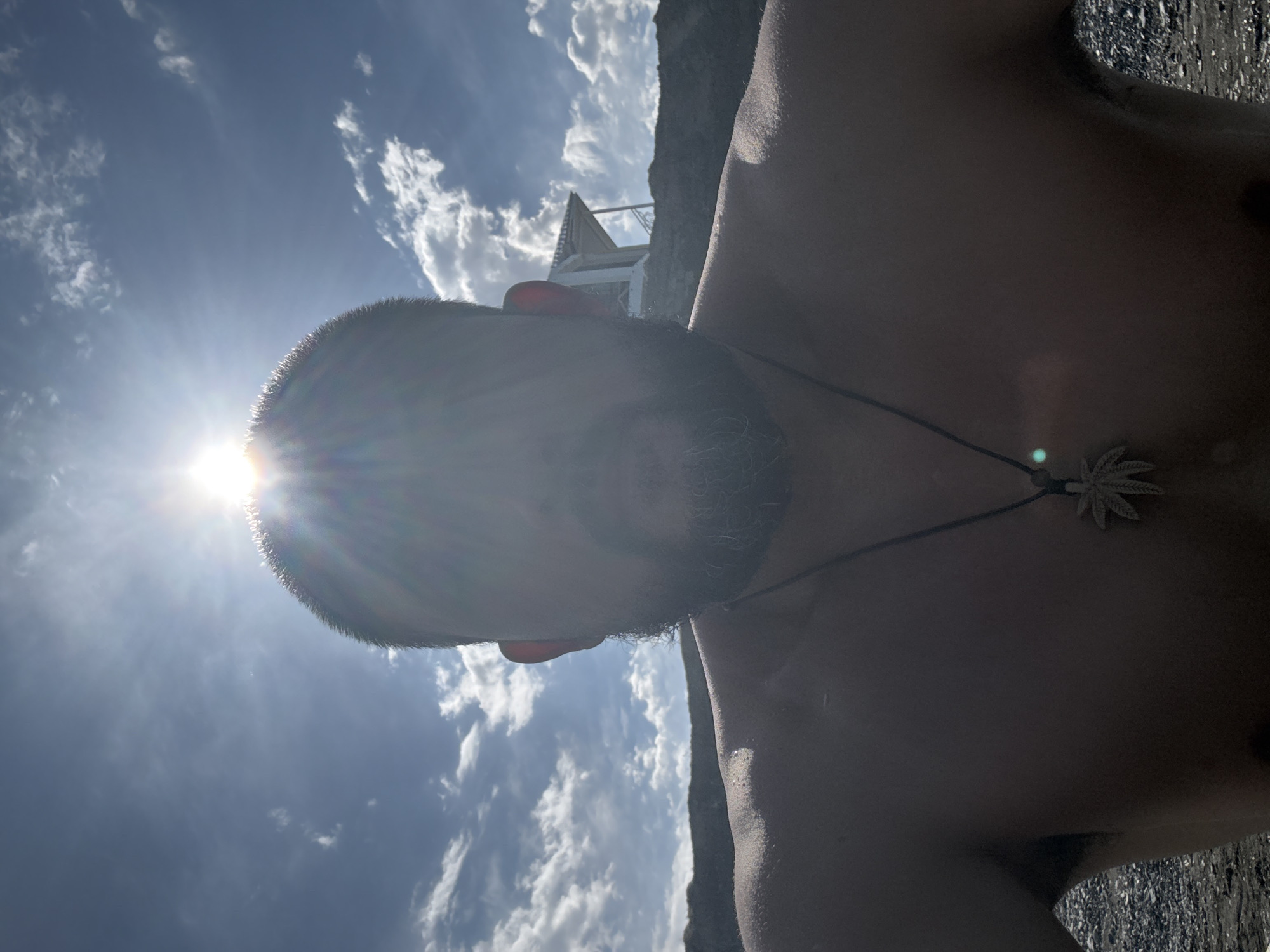 The image is a close-up, waist-up, selfie-style portrait of a man with a beard, taken outdoors against a bright sky. The sun is positioned directly behind the man's head, creating a lens flare effect that obscures his face and casts his upper body in silhouette. He is shirtless, wearing a necklace with a leaf-shaped pendant. In the background, a small, white, open-air structure with a peaked roof is visible on a rocky incline, silhouetted against a blue sky with scattered white clouds. The ground in the foreground appears to be a dark, pebbly surface, possibly a beach. The overall lighting suggests a sunny day. The location is not identifiable as Ақши, Kazakhstan, from the visual information provided in the image.