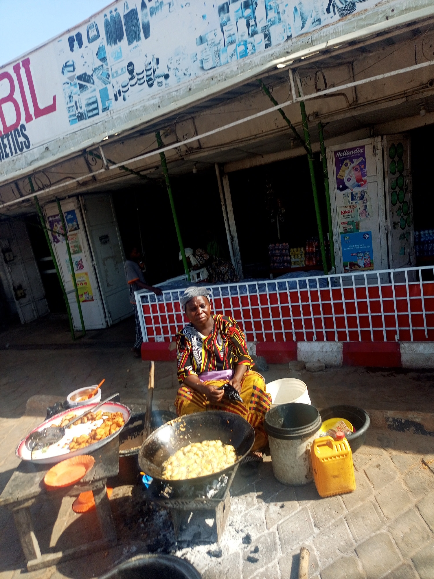 The image captures an outdoor street food scene in Tempure, Nigeria, under bright, clear daylight. An older woman, likely a vendor, sits on a low stool or directly on the paved ground. She wears a vibrant, multi-colored patterned dress, typical of traditional African attire, and has a neutral, possibly weary, expression.
In front of her, a makeshift cooking station is set up. One large black wok contains light-colored food, possibly fried plantains or potatoes, actively cooking over a small, glowing charcoal or wood fire supported by a rudimentary metal stand. Adjacent to it, a metal serving tray holds a quantity of similar-looking cooked food. Other items visible around her include a small white bowl with a red spoon, an orange plastic plate, a bright yellow jerrycan (likely for cooking oil), a white plastic bucket, and a black basin.
The cooking area is on a paved street, in front of a row of shop stalls, most of which appear closed or partially open. A low red and white patterned barrier runs along the front of the shops. Above the stalls, a large billboard reads "BIL COSMETICS" and features various cosmetic product illustrations. Advertisements for "Hollandia" (mentioning "fresh yoghurt") and "KING'S" are visible on posters affixed to the shop doors. Strong shadows indicate a sunny day, suggesting morning or early afternoon.