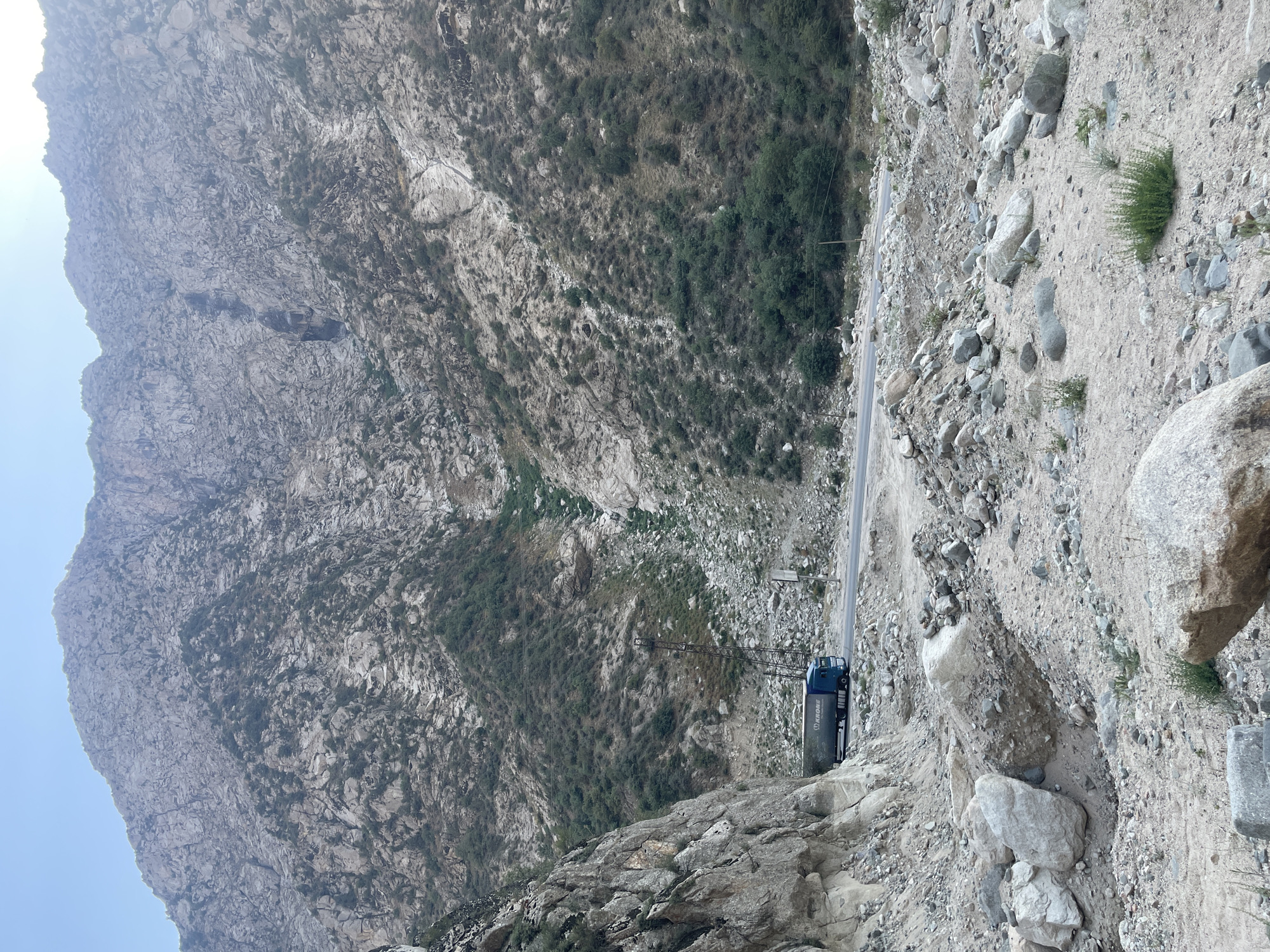 The image depicts a mountain landscape in Khodzha-Obigarm, Tajikistan, with a paved road winding through a rocky, arid terrain. A blue semi-trailer truck with "KRONE" branding is driving on the road. The mountains are steep and rugged, with sparse vegetation and evidence of rockfalls. The sky is clear and bright. The foreground is filled with rocks and debris, suggesting a natural or possibly man-made alteration of the landscape. Power lines are visible stretching across the mountainside.