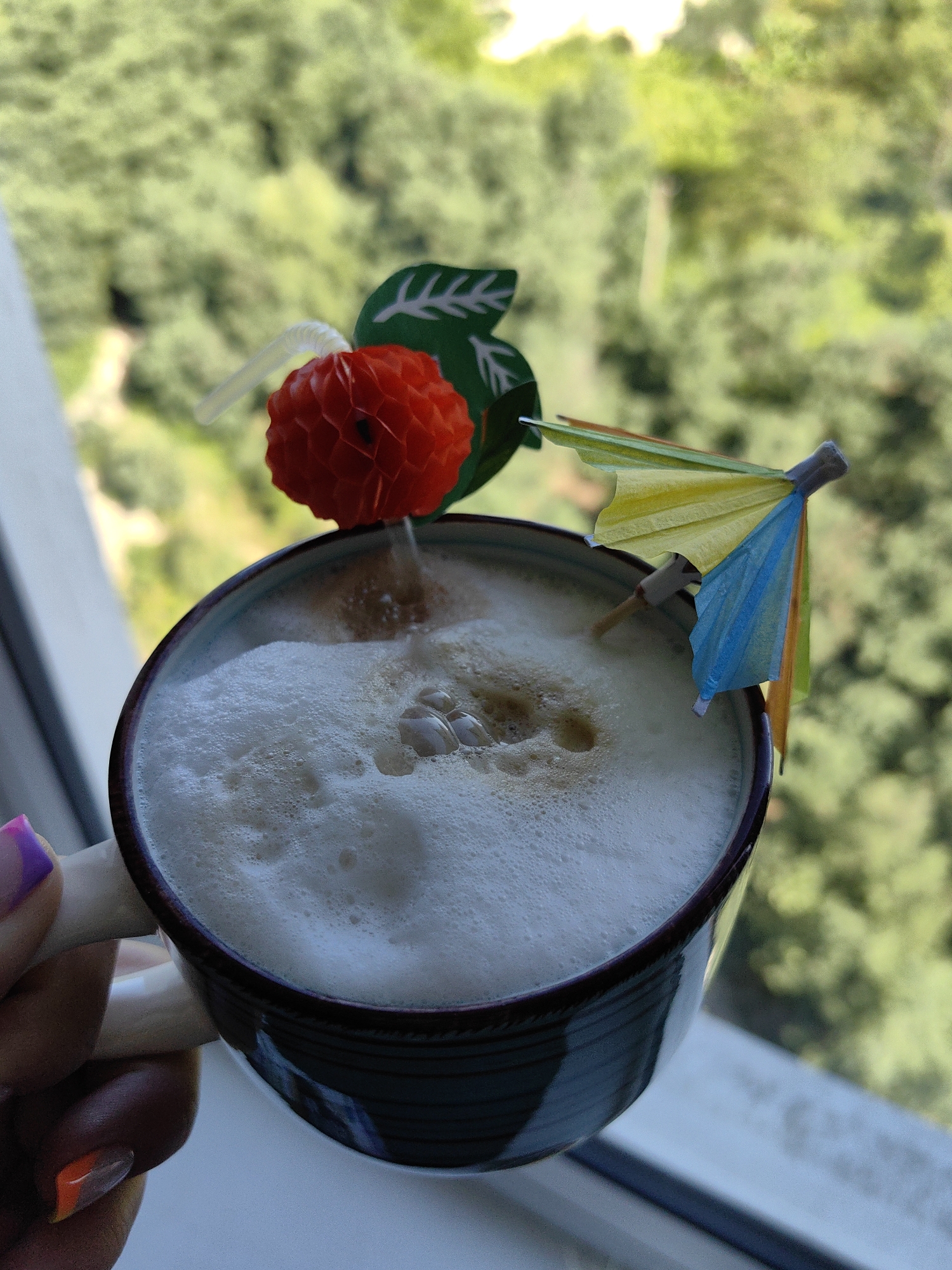A close-up, high-angle shot shows a dark blue ceramic mug filled with frothy coffee, garnished with a red honeycomb ball on a straw and a small colorful umbrella. The mug is held by a person whose hand with brightly painted nails is visible on the left side of the frame. The background is a blurred view of lush green trees, suggesting an outdoor setting, possibly a balcony or window overlooking foliage. The scene is likely taking place in Sochi, Russia, given the abundant greenery and the typical summer weather suggested by the light.