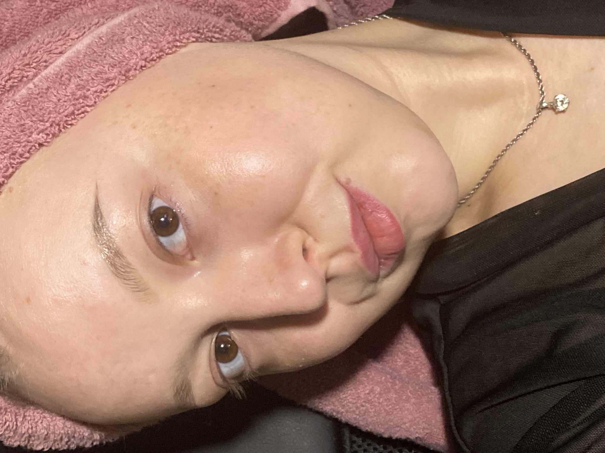 A fair-skinned person with light brown eyes and freckles is lying on their back. They have a pink towel wrapped around their head. A silver chain necklace with a diamond pendant is visible on their neck. They are wearing a black mesh top. The location and city of Chaikovsky, Russia are not evident in this image.