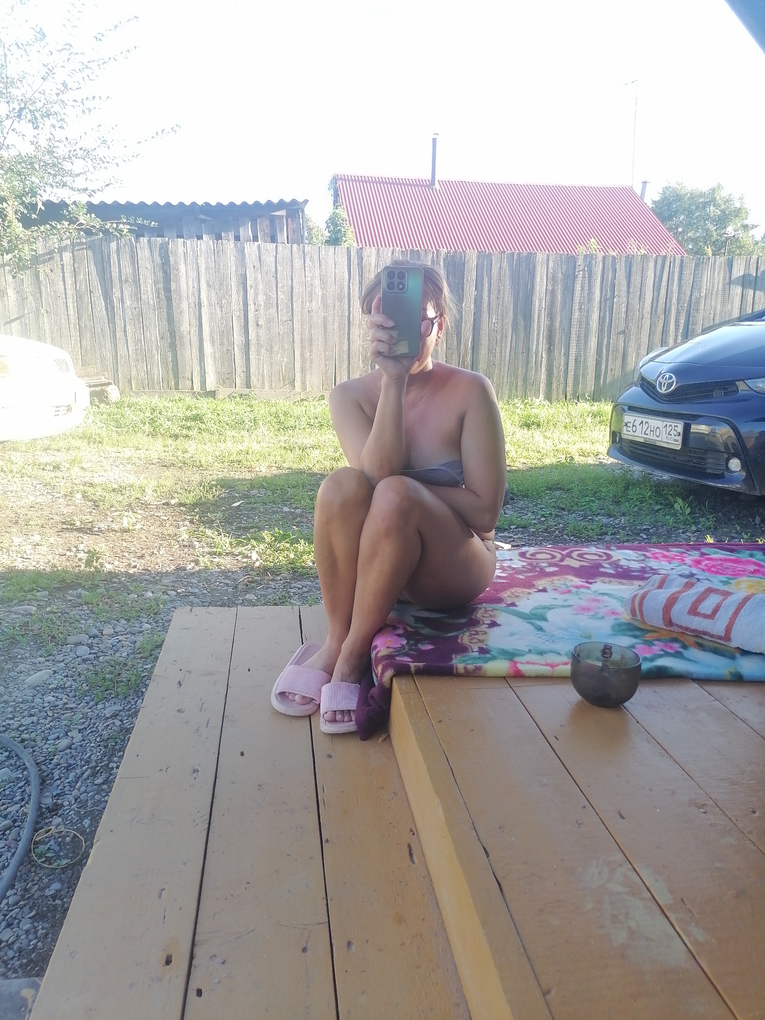 A woman is sitting on a wooden platform, facing left, holding a phone in front of her face, obscuring it. She is wearing pink slippers. A colorful blanket is spread out to her right, with a towel and a small dark bowl on top of it. Behind her, a wooden fence runs horizontally, with a red-roofed building visible beyond it. A dark blue car is parked to her right. The ground is covered in gravel and grass. The scene appears to be outdoors in a rural or suburban setting.
